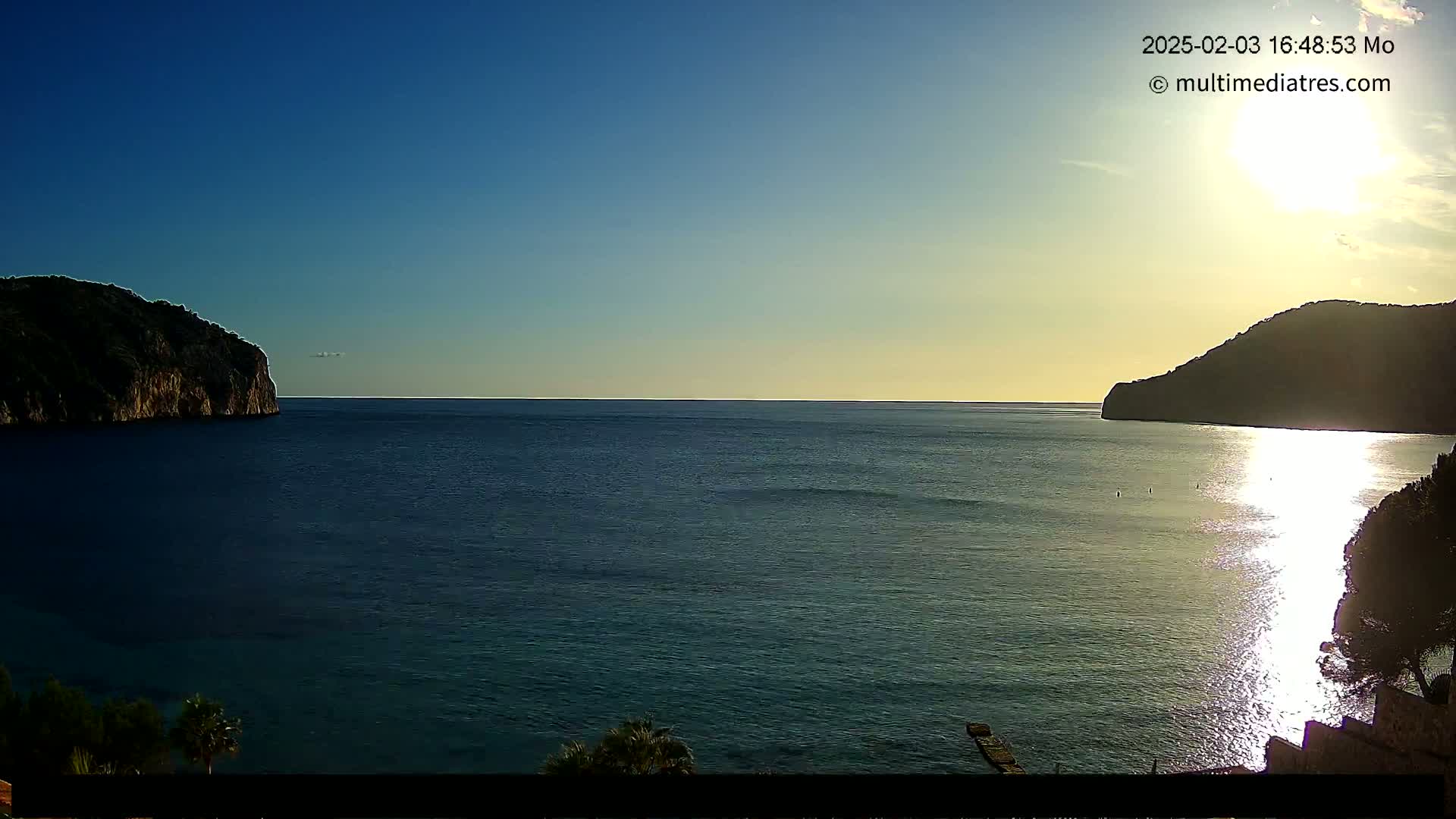 A calm sea is seen at sunset between two dark, rocky headlands under a clear blue sky.