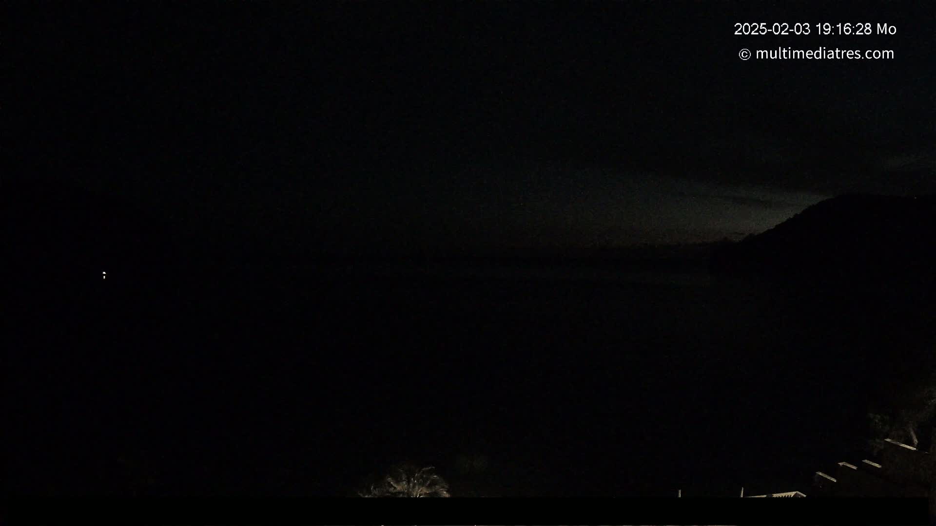 A dark nighttime scene shows a body of water in the distance, a dark hillside to the right, and a few faint lights in the bottom left and right corners.