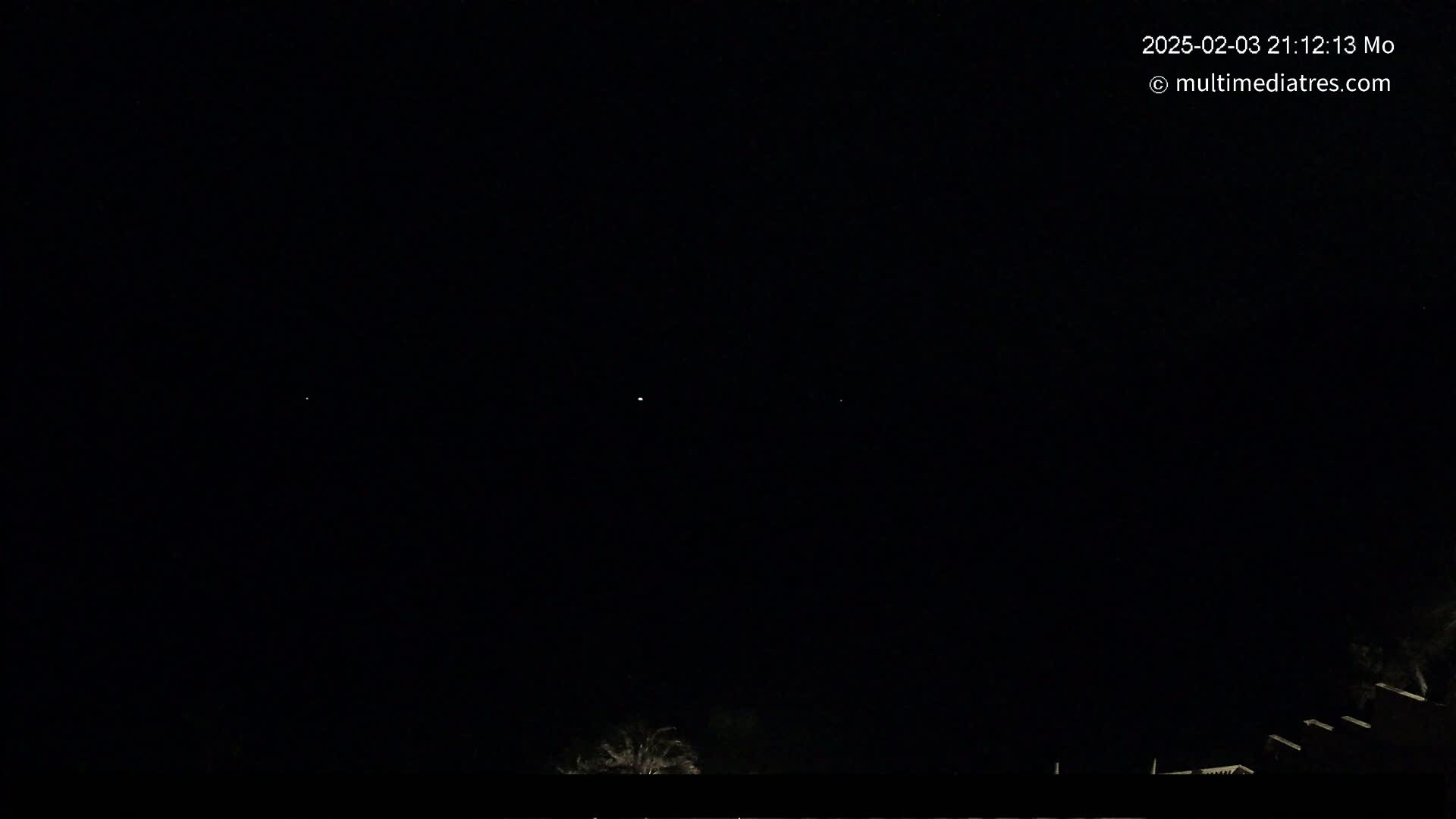 A dark nighttime sky with two faint lights visible and the silhouettes of buildings and trees at the bottom.