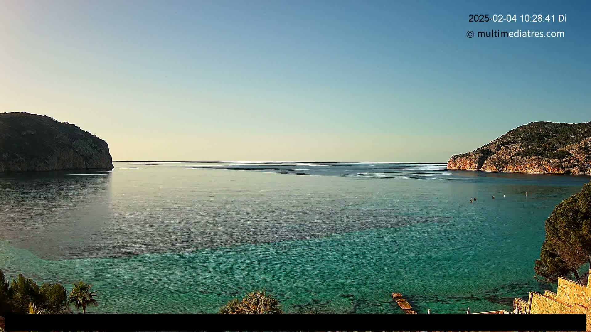 A calm, clear day reveals a tranquil bay of teal water between two rocky hillsides, with a few people visible in the distance.