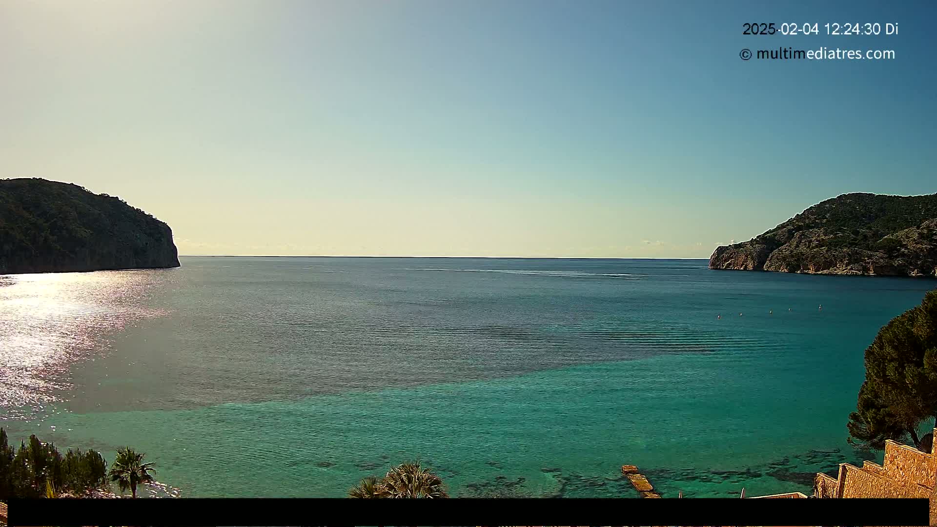 A calm, sunny day reveals a tranquil bay with clear, turquoise water, nestled between two dark, rocky hillsides.