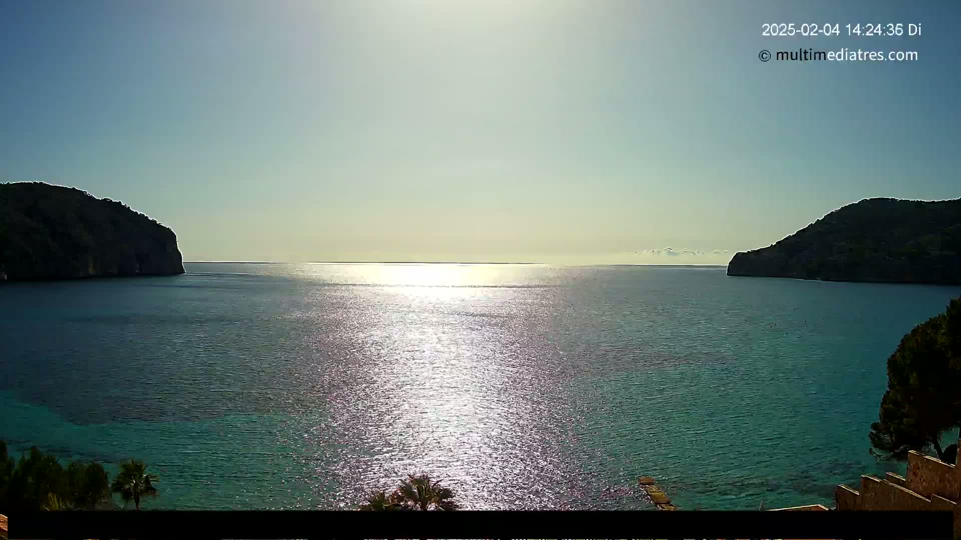 A calm, sunny day reveals a bay of clear turquoise water between two dark, rocky hills.