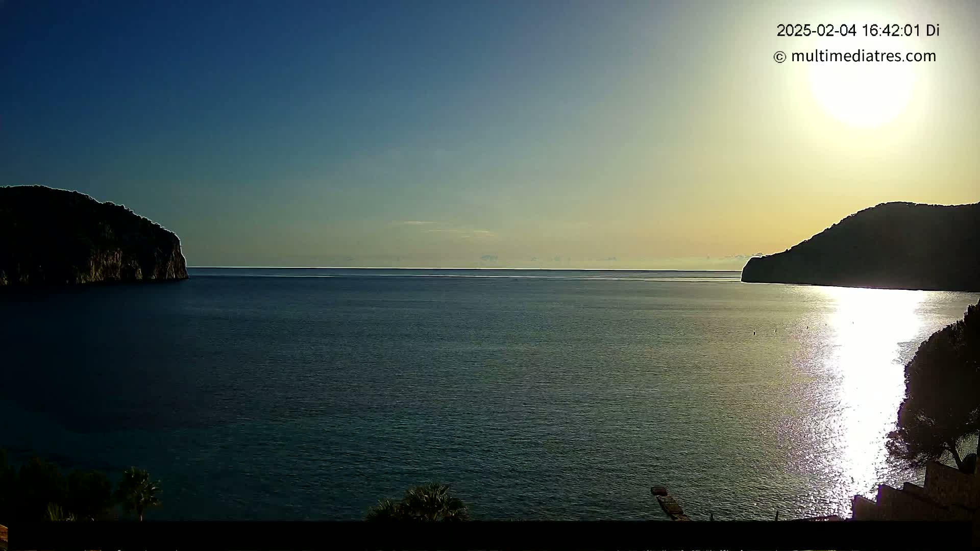 A calm sea is seen between two dark, rocky hillsides under a clear, bright sky with the sun shining on the water.