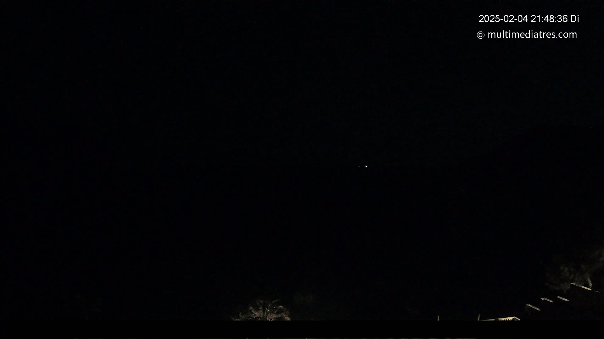 A nighttime view shows a dark sky with a few faint lights and the tops of dark trees or buildings at the bottom.