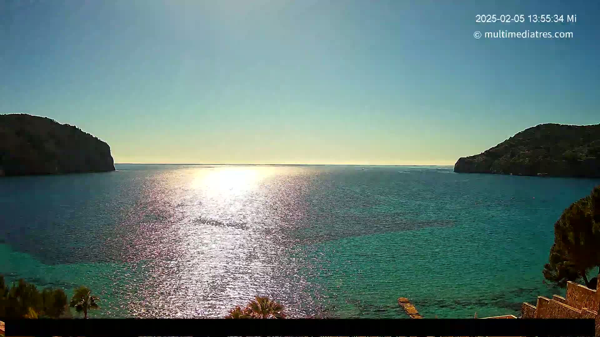 A calm, sunlit sea is nestled between two dark, rocky hills, under a clear blue sky.