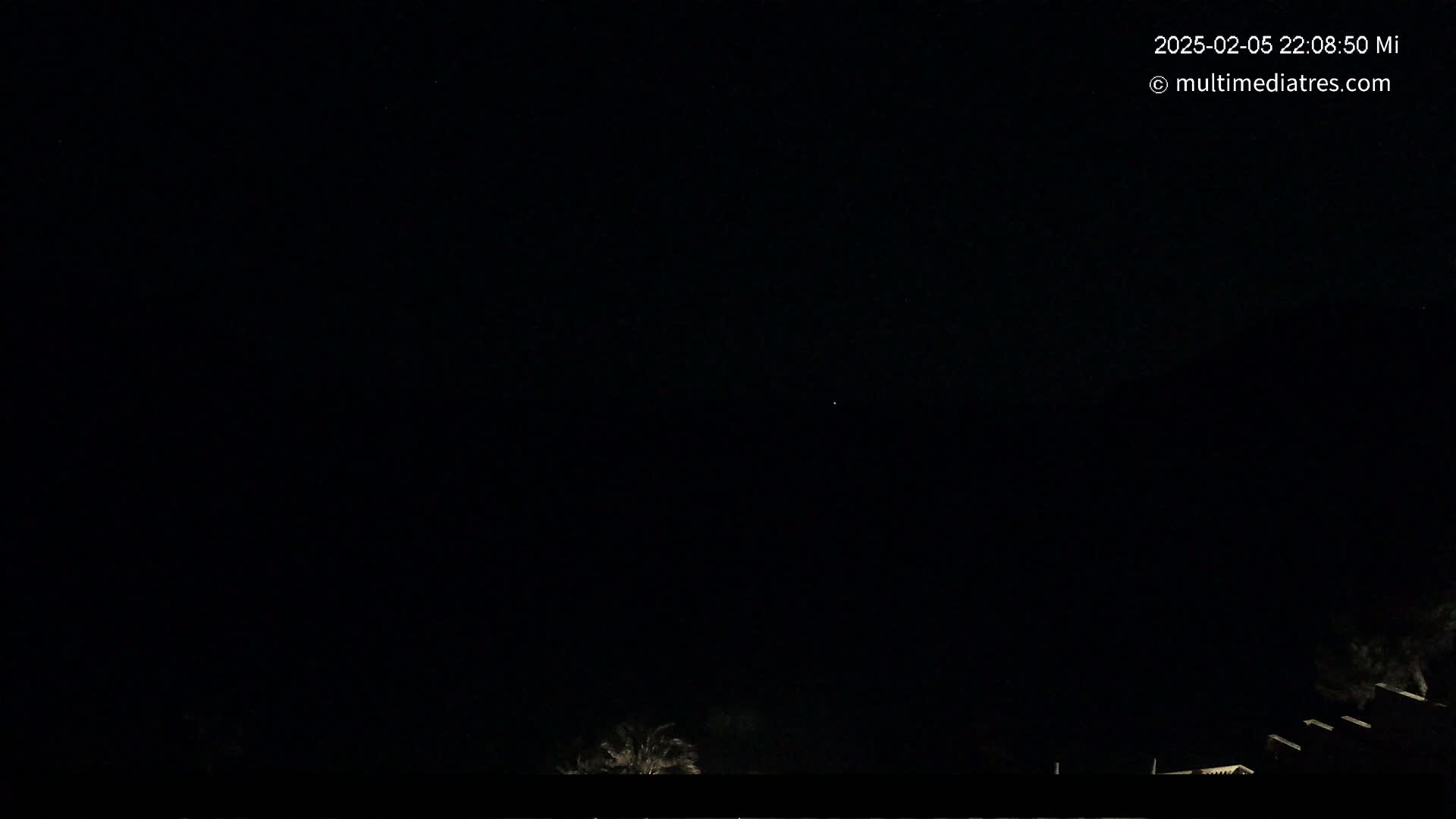 A nighttime, outdoor scene shows a mostly dark sky with a few faint stars above a low silhouette of buildings and trees.