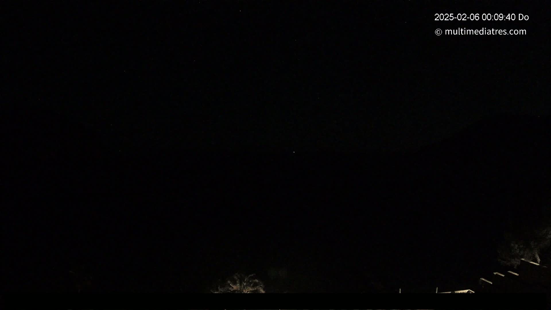 A nighttime, outdoor scene shows a mostly clear, dark sky with a few stars visible above a dark silhouette of trees and structures at the bottom of the frame.