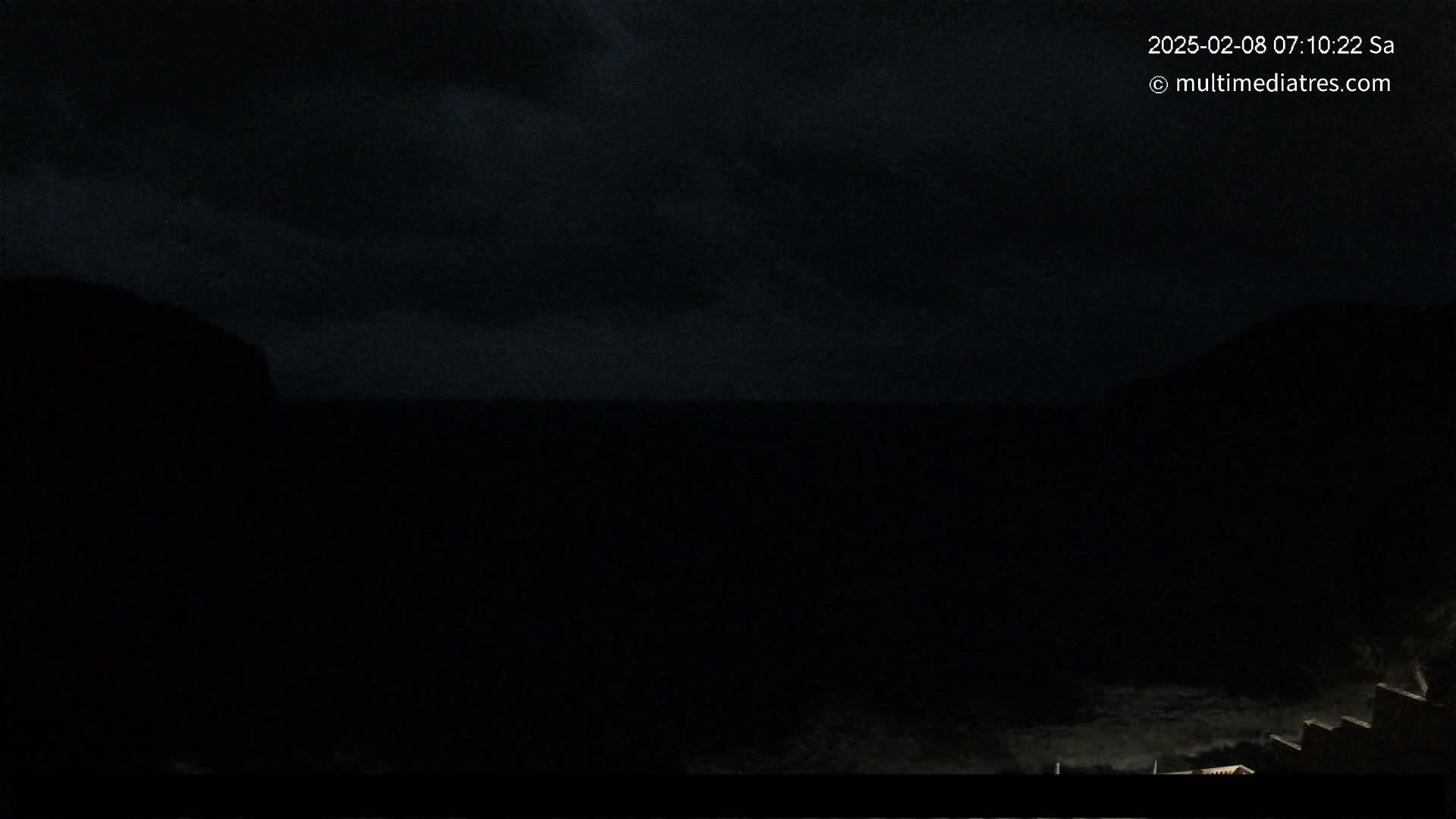 A dark, nighttime coastal scene shows a body of water between two silhouetted landmasses under a cloudy sky.