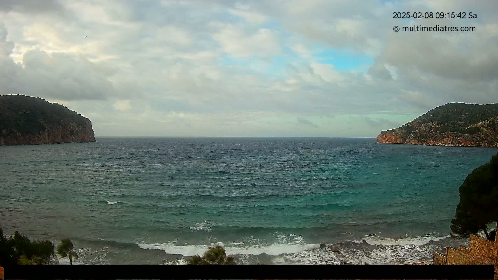A partly cloudy day shows a wide expanse of teal ocean with small waves lapping a shore between two dark, rocky hills.