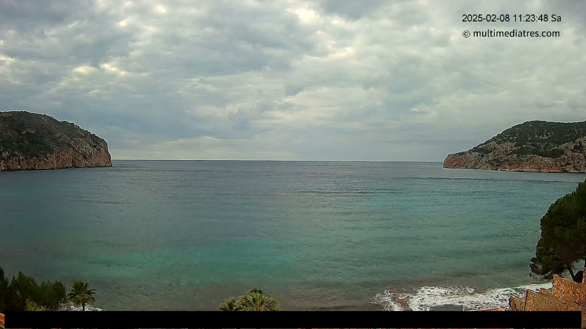 A calm, teal-colored bay is nestled between two rocky, tree-covered hills under a cloudy sky.