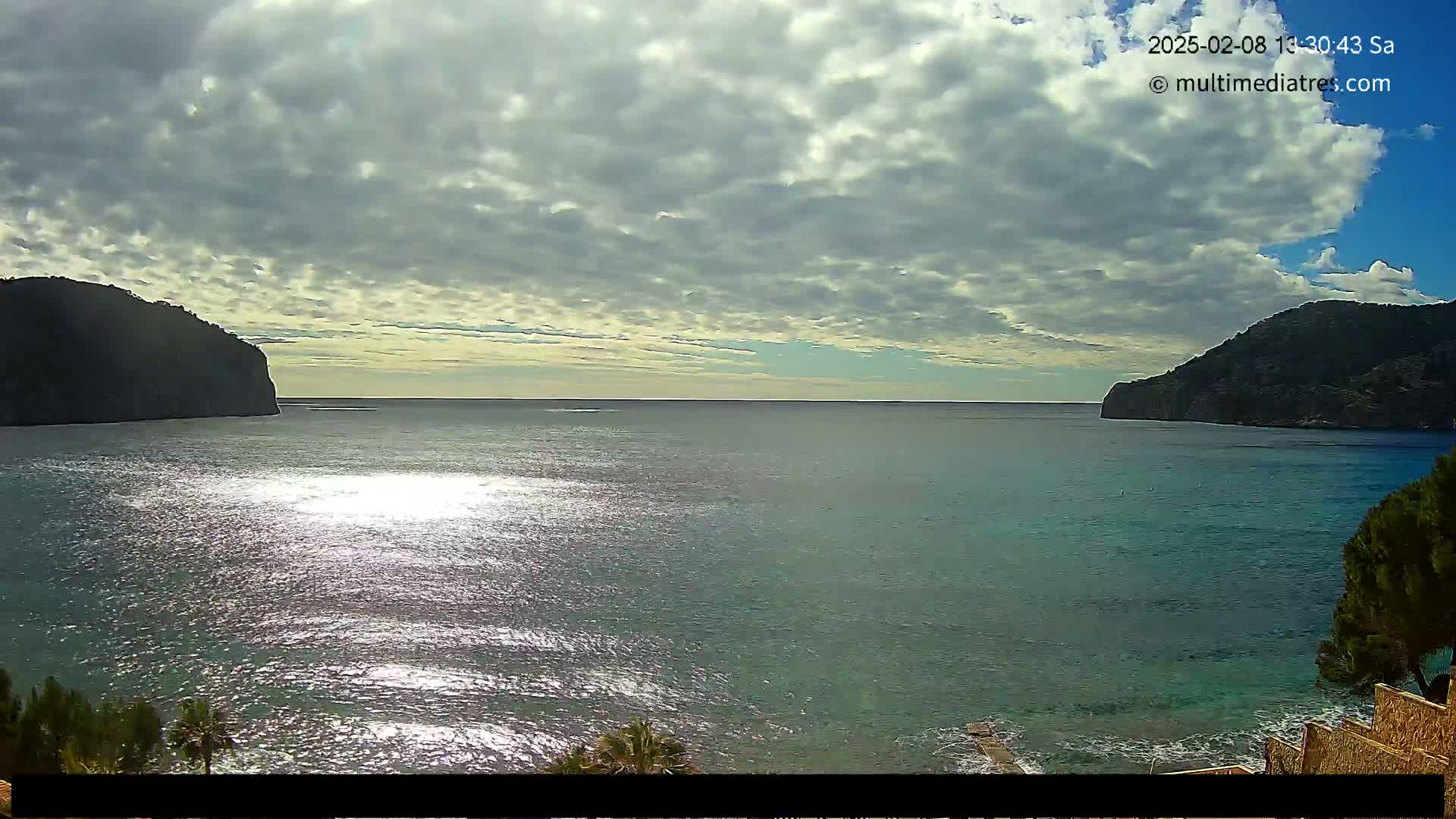 A partly cloudy day reveals a calm, sparkling body of water nestled between two dark, rocky hills.