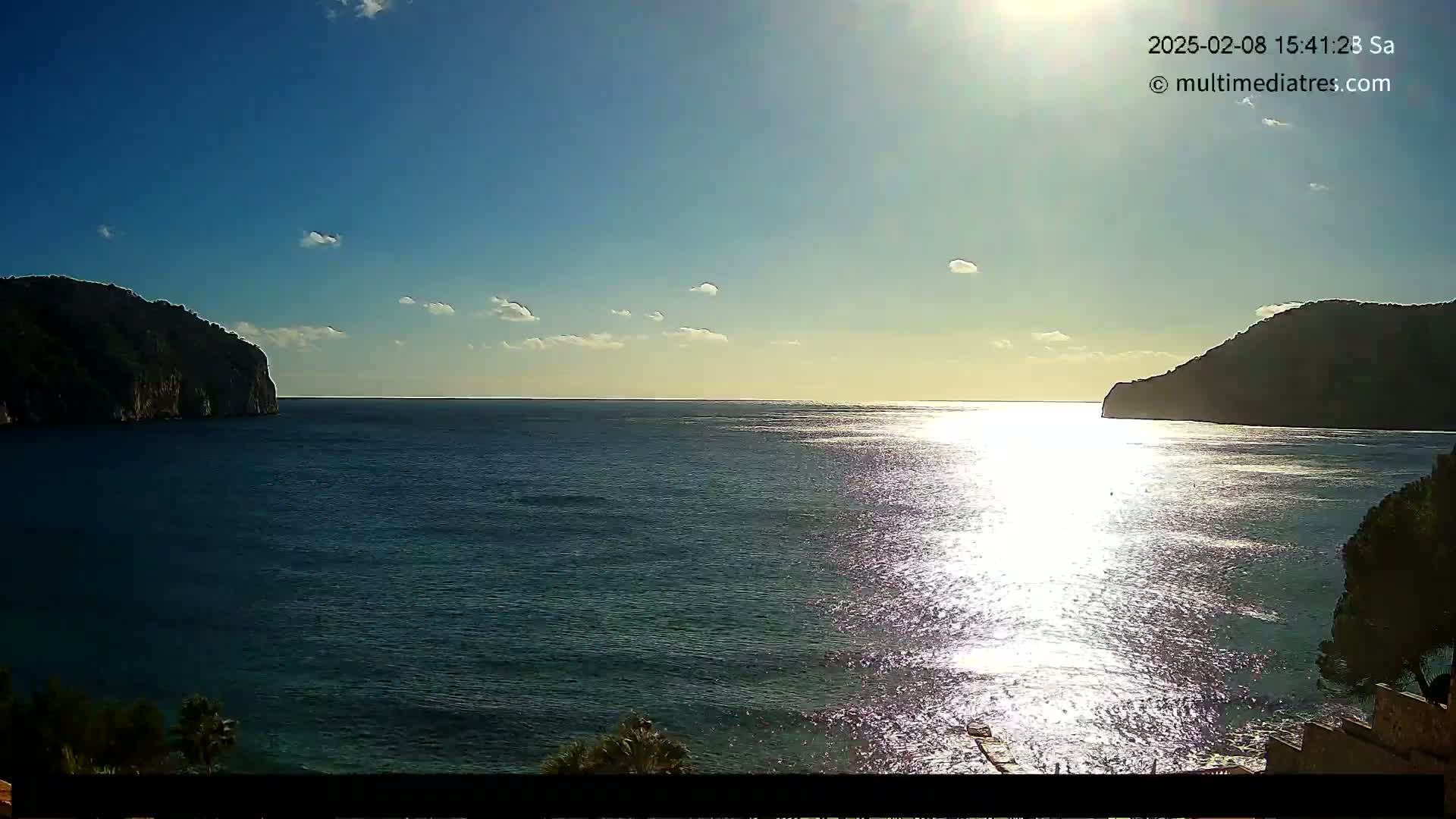 A sunny day shows a calm sea between two dark hills, with the sun's reflection sparkling on the water.