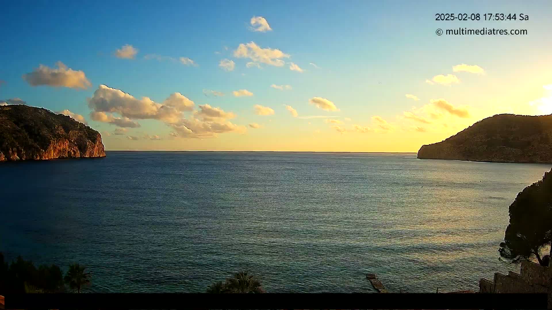 A calm sea at sunset is nestled between two rocky hills under a partly cloudy sky.