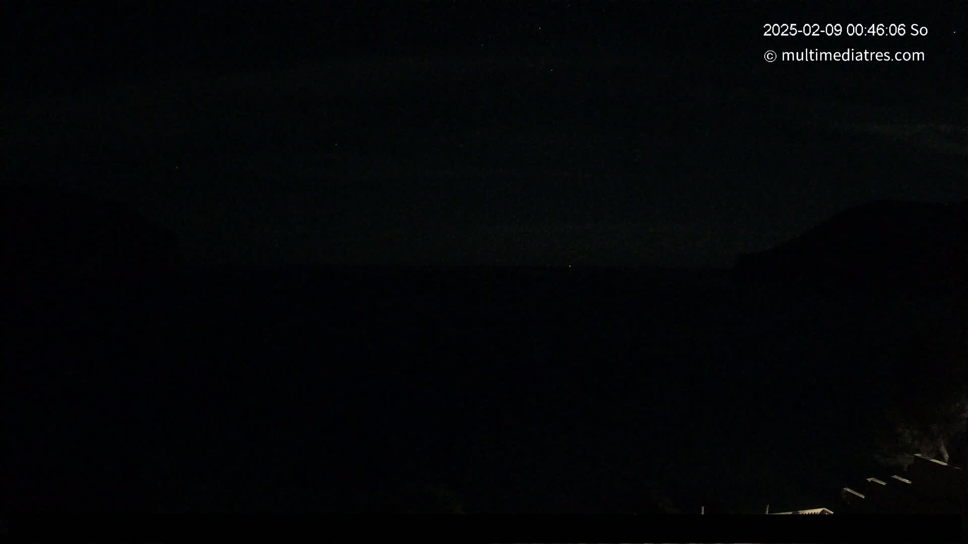 A dark, nighttime scene shows a mostly clear sky above a dark landscape with a few faint lights visible near the bottom.