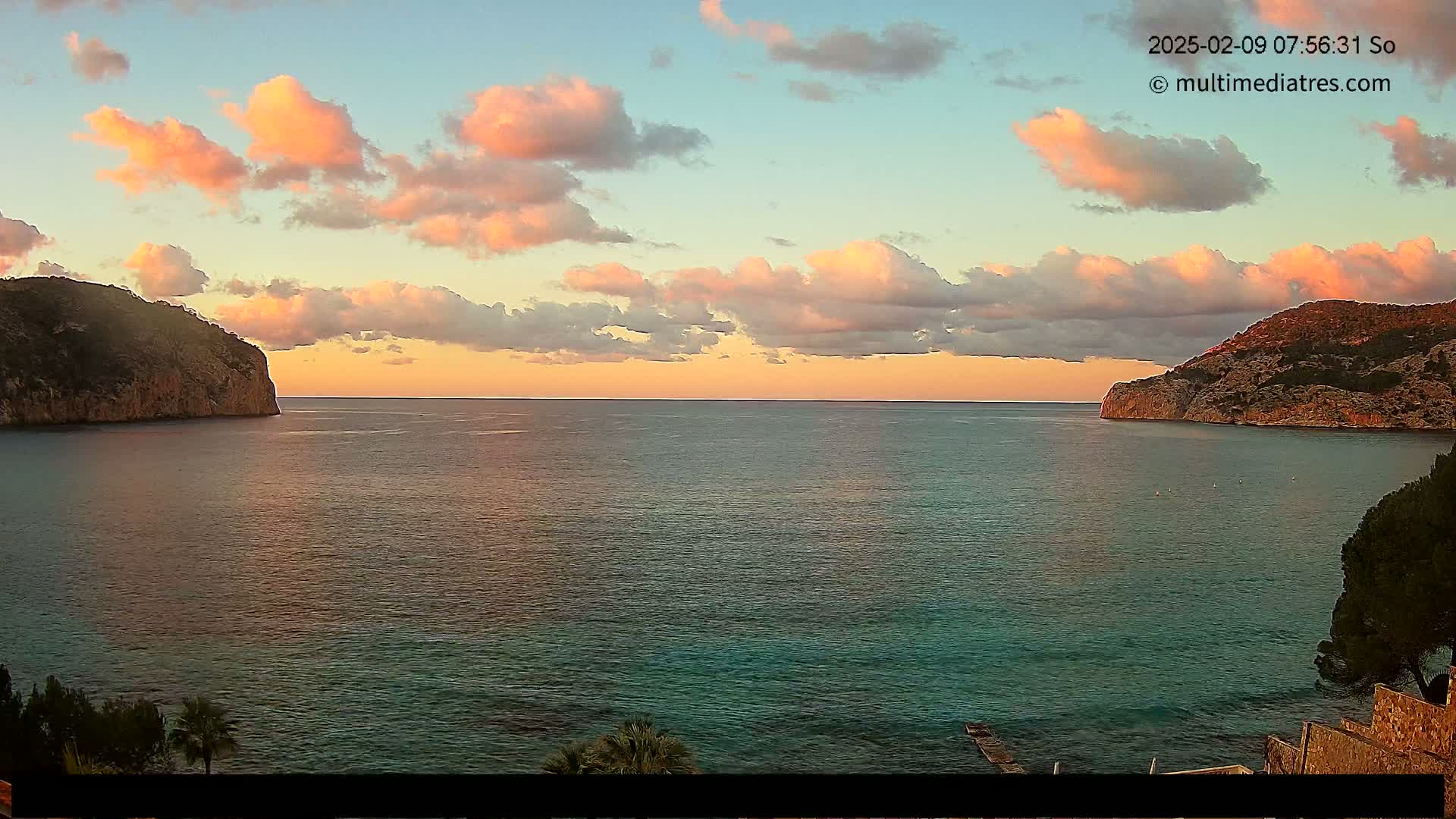 A calm sea at sunset is nestled between two rocky cliffs under a sky filled with soft pink and orange clouds.