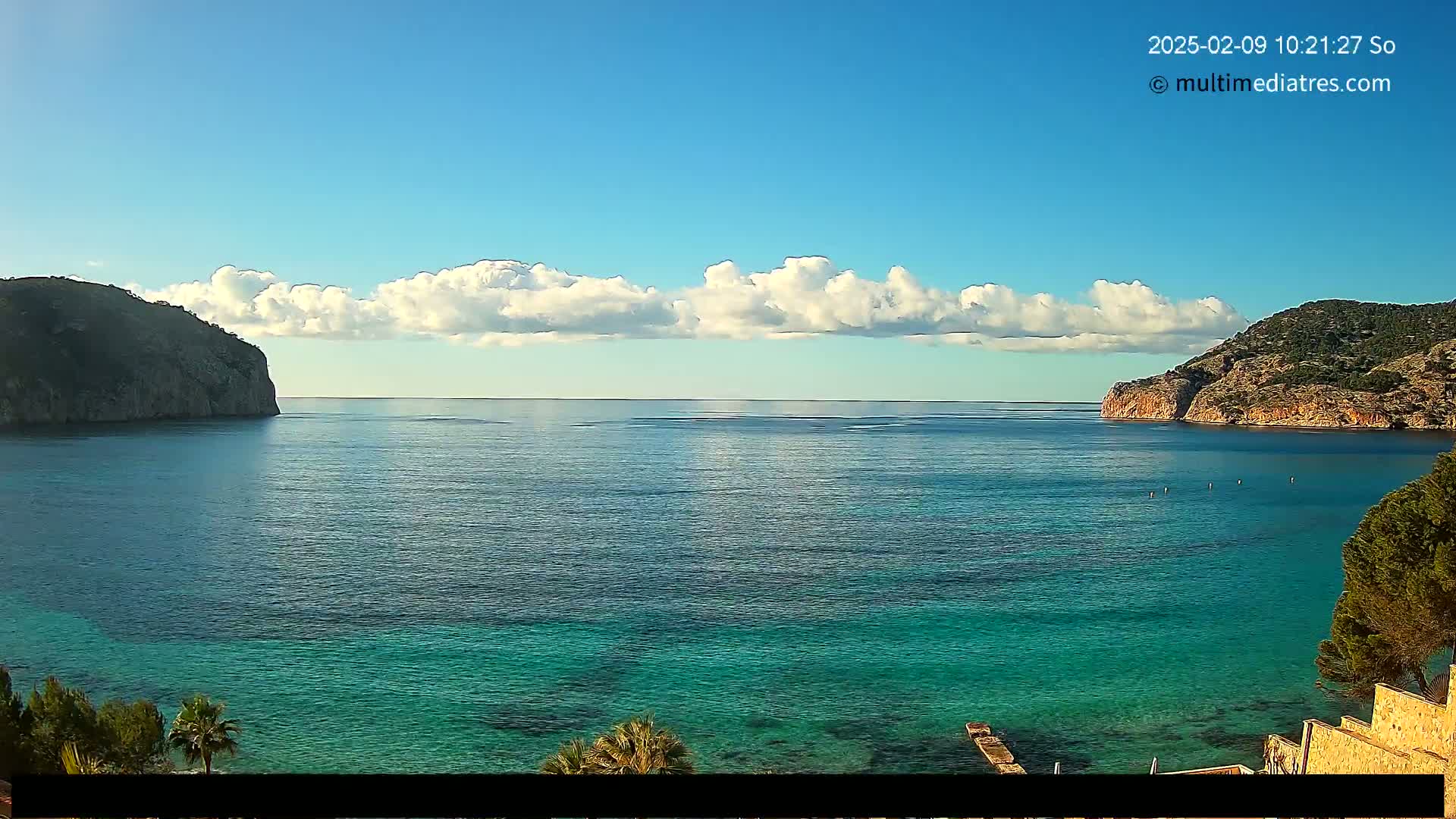 A tranquil coastal scene shows a calm, turquoise sea between two rocky hills under a mostly clear sky with a band of fluffy white clouds.
