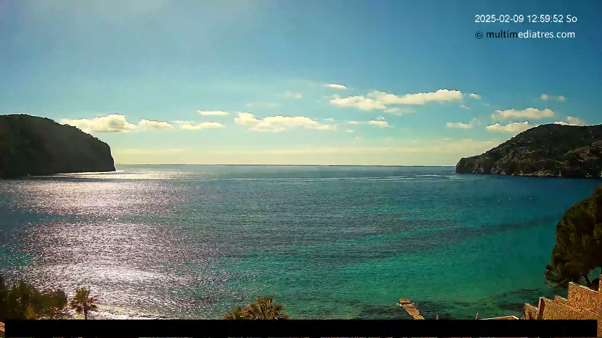 A tranquil cove of clear, teal water is nestled between two dark, verdant hills under a mostly sunny sky with scattered clouds.