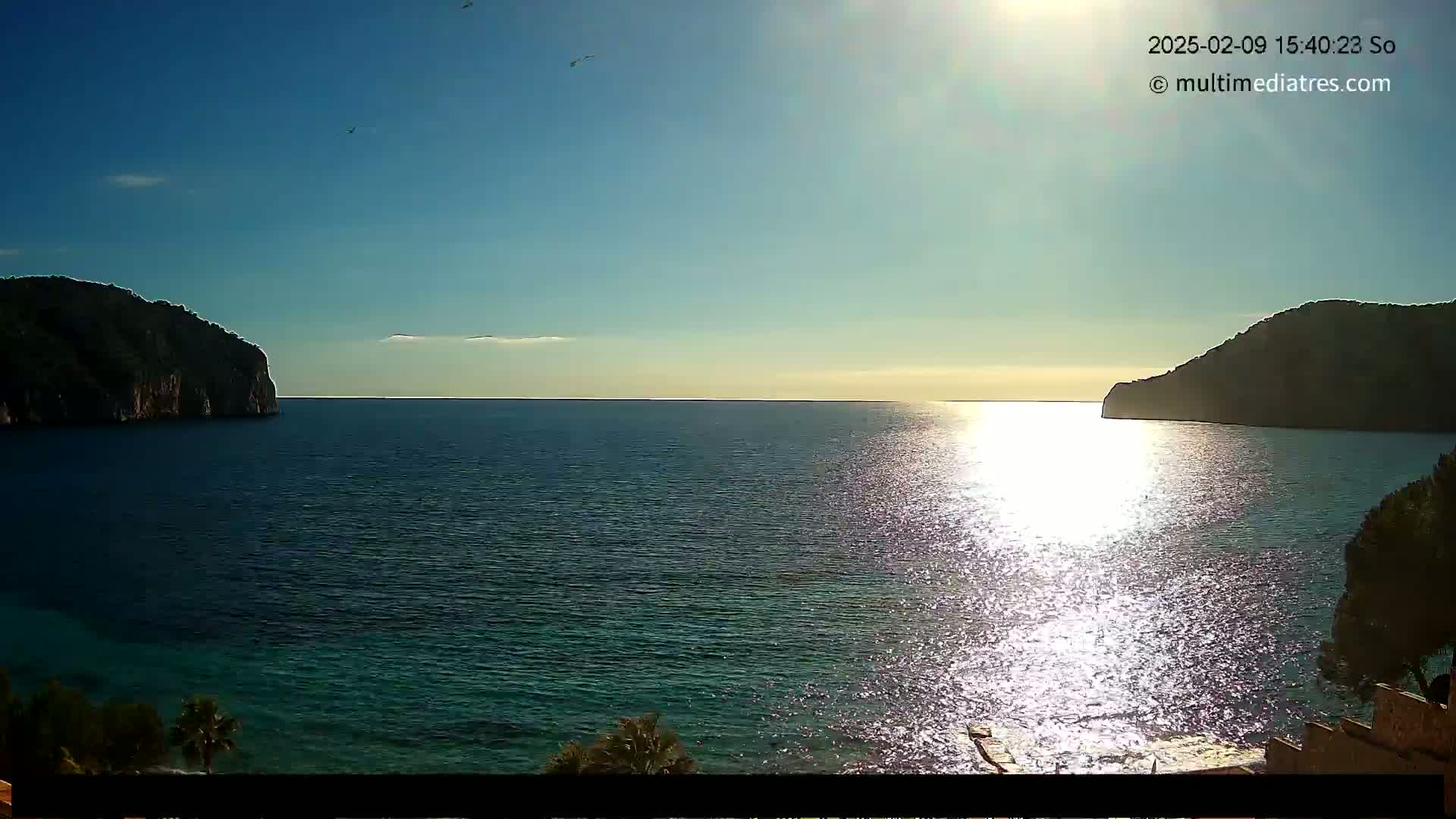 A calm, sunlit sea is nestled between two dark, rocky hills under a clear blue sky.
