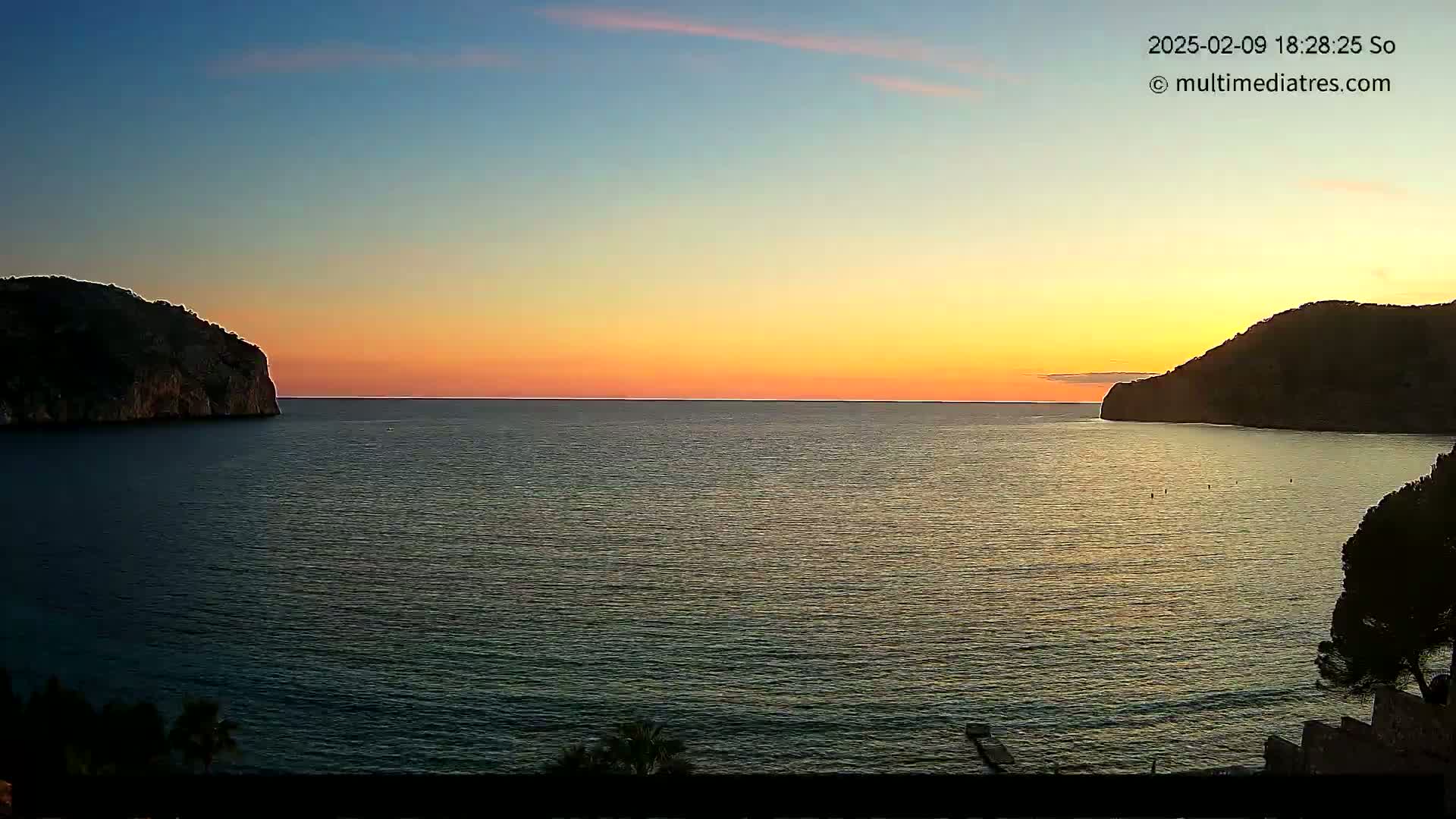 A calm sea at sunset is nestled between two dark, rocky hillsides.