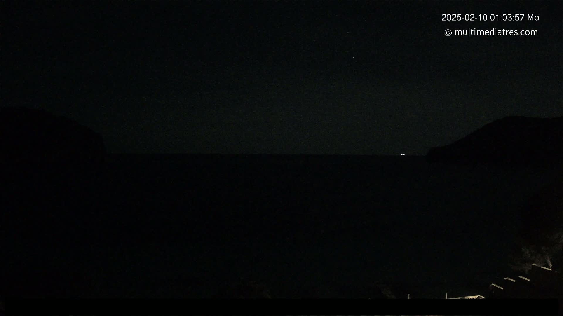 A dark, nighttime view of a calm body of water between two dark landmasses, with a single small light visible on the horizon.