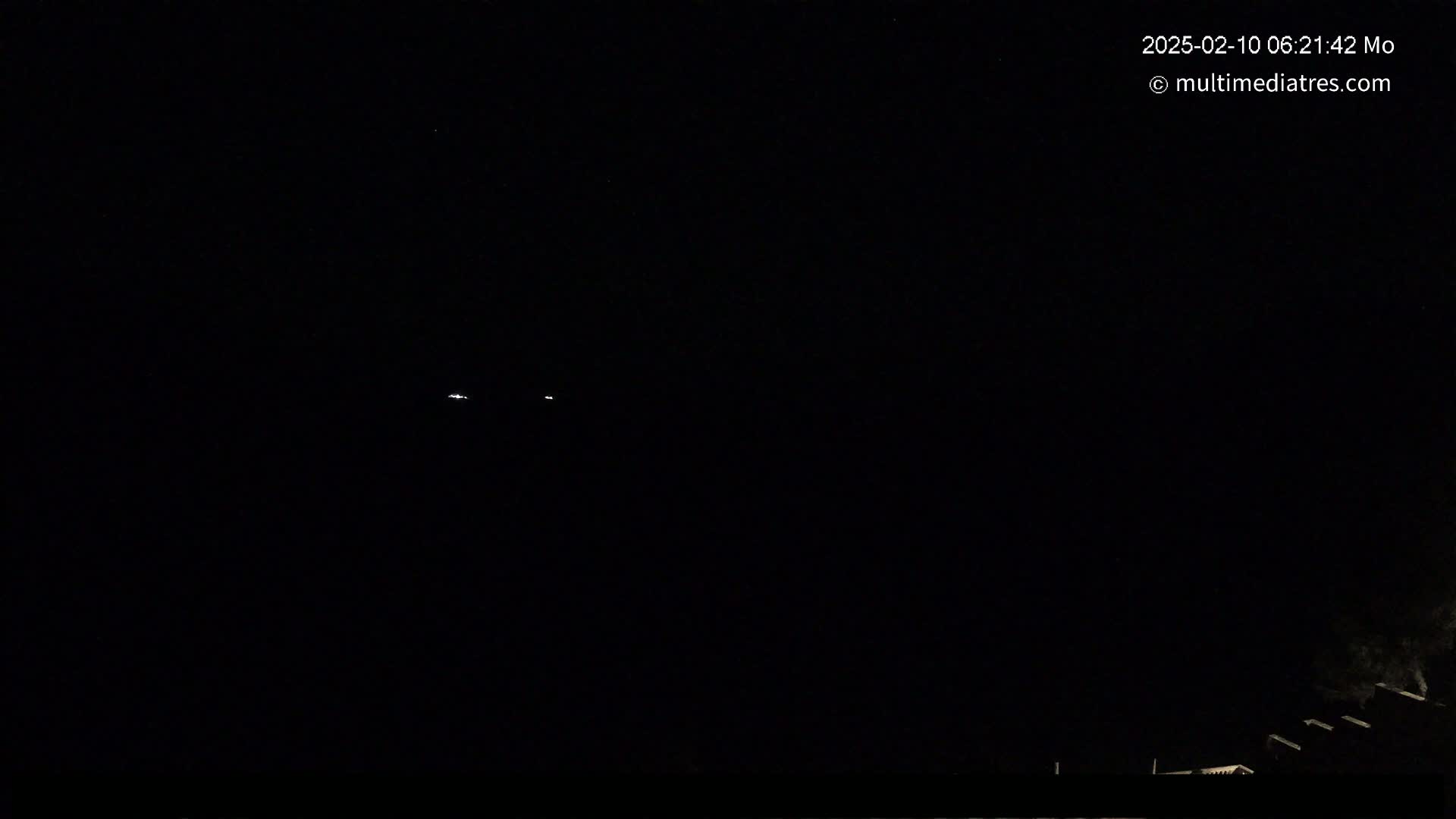 A nighttime, dark scene shows a few faint lights in the distance and a dark structure at the bottom of the frame.