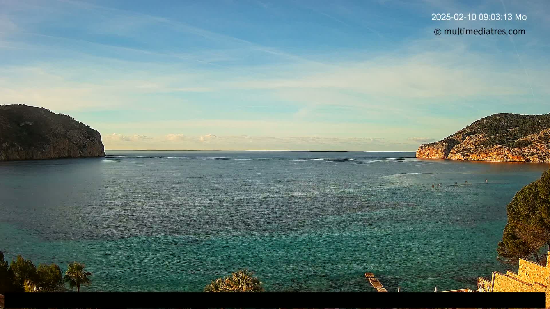 A calm, clear day reveals a tranquil bay with teal water, two rocky headlands, and several people paddleboarding in the distance.