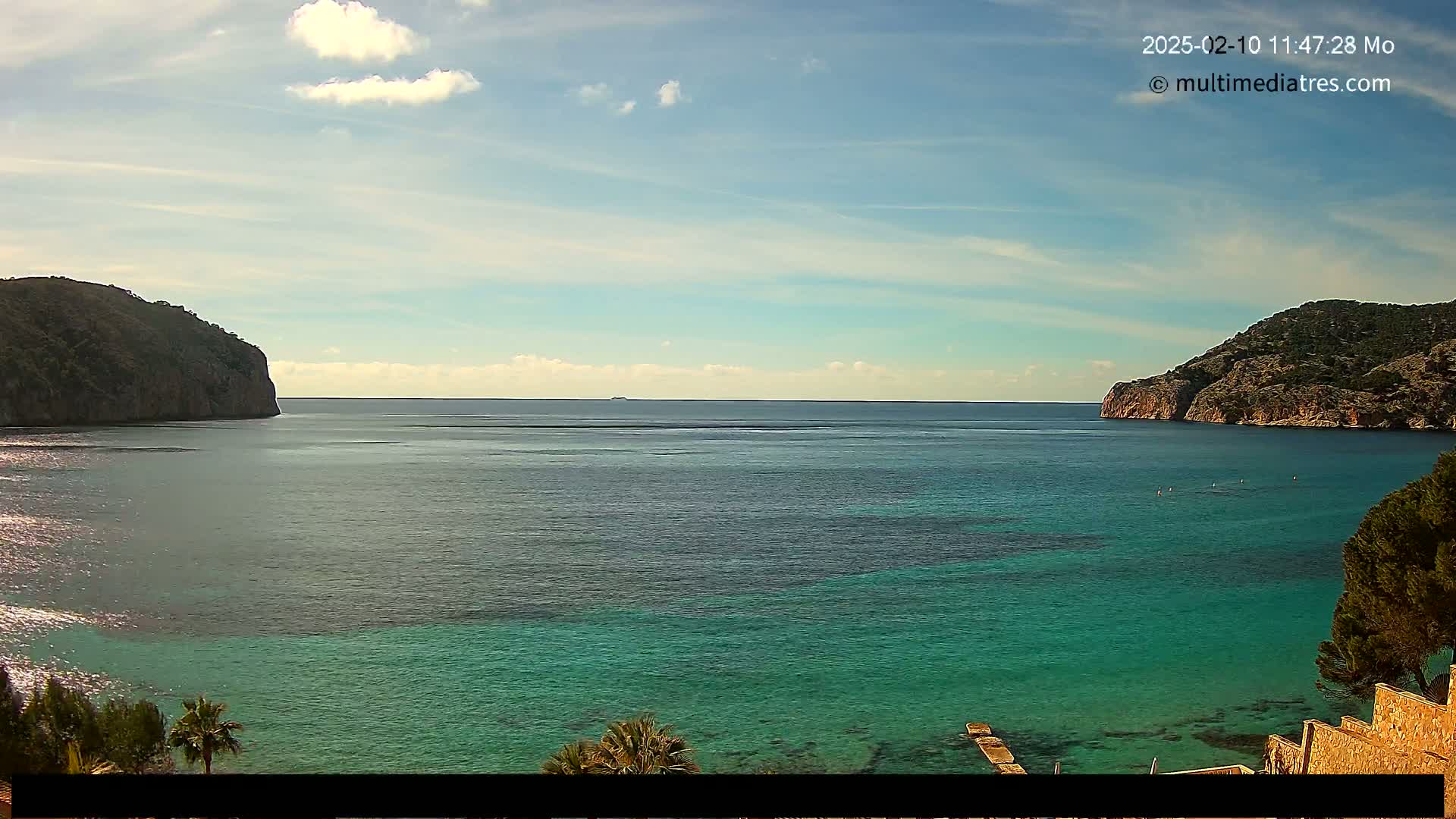 A calm, clear day reveals a tranquil bay of turquoise water nestled between two rocky hillsides.