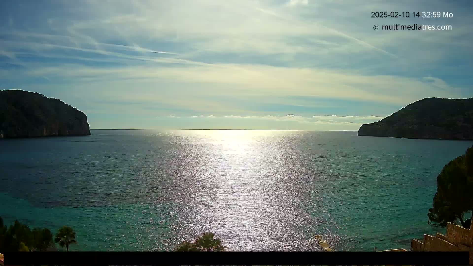 A calm, sunlit sea is nestled between two dark, rocky hills under a mostly clear sky.
