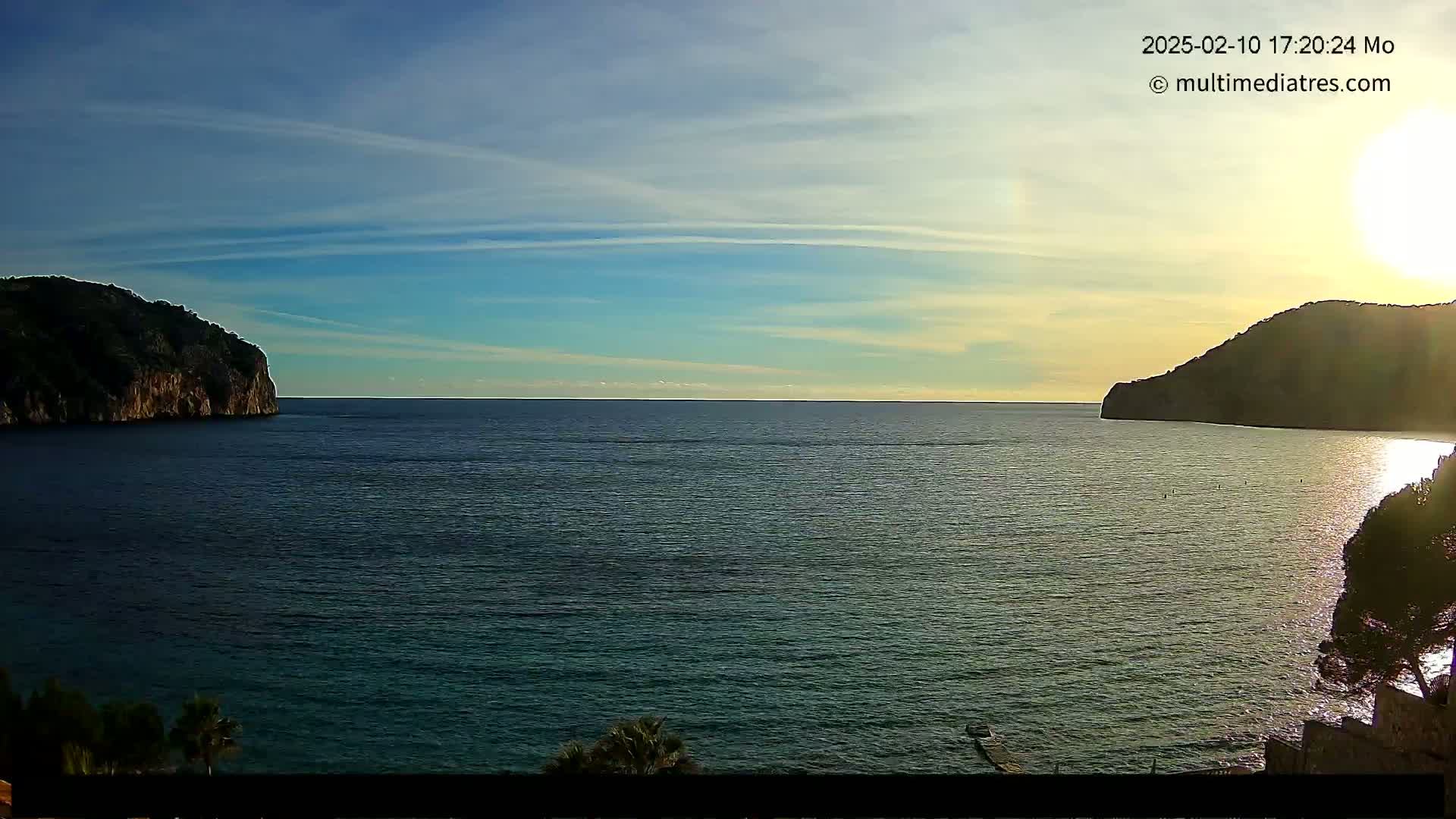 A calm sea is nestled between two dark, rocky hills under a mostly clear, sunset sky.