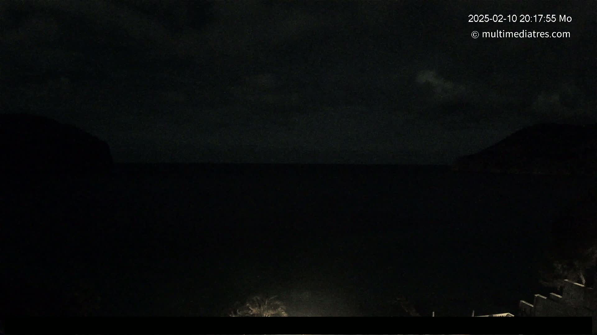 A nighttime, dark view of a calm body of water between two silhouetted hills under a cloudy sky.