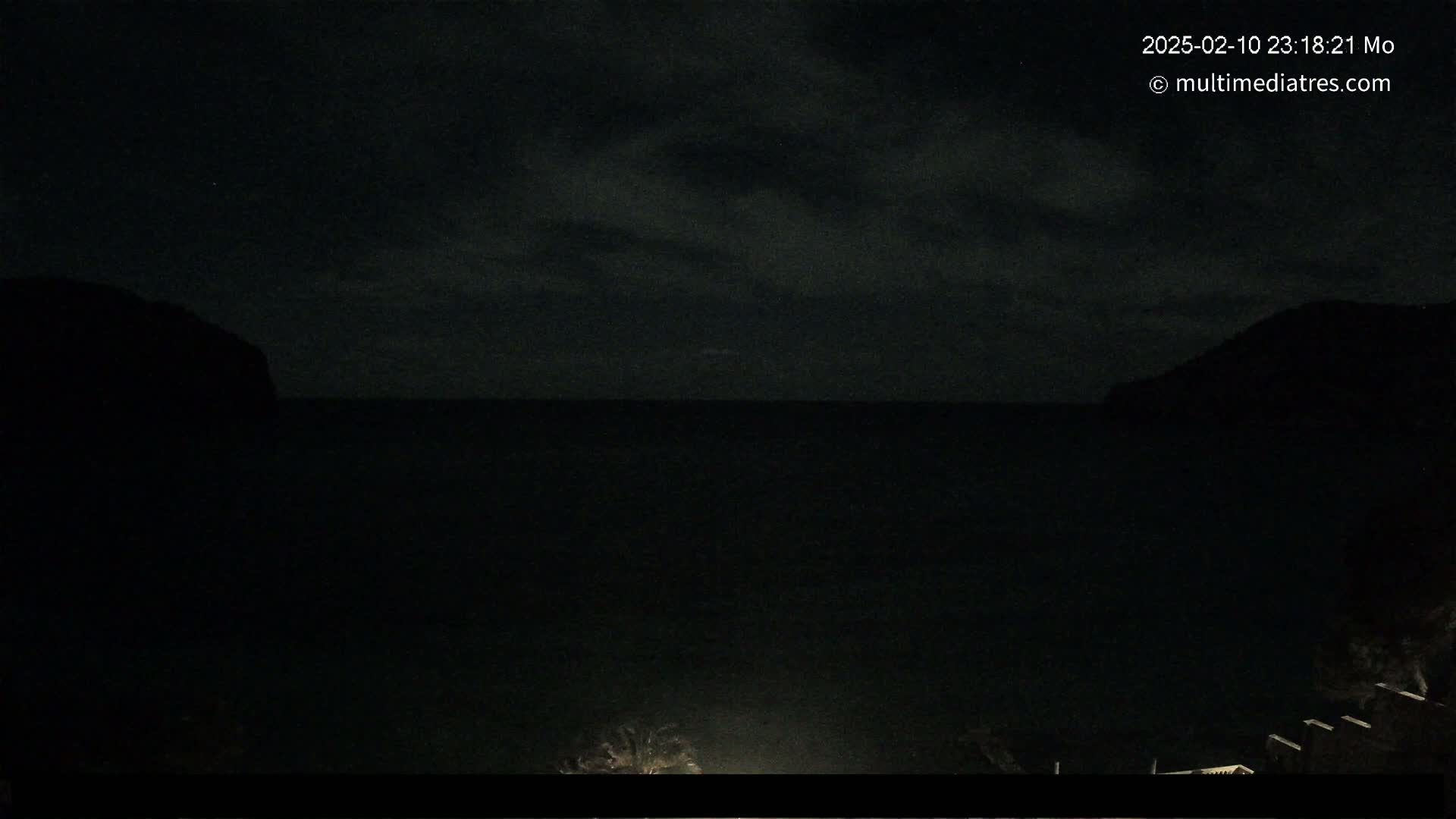 A nighttime view of dark, calm water between two silhouetted hills under a cloudy sky.