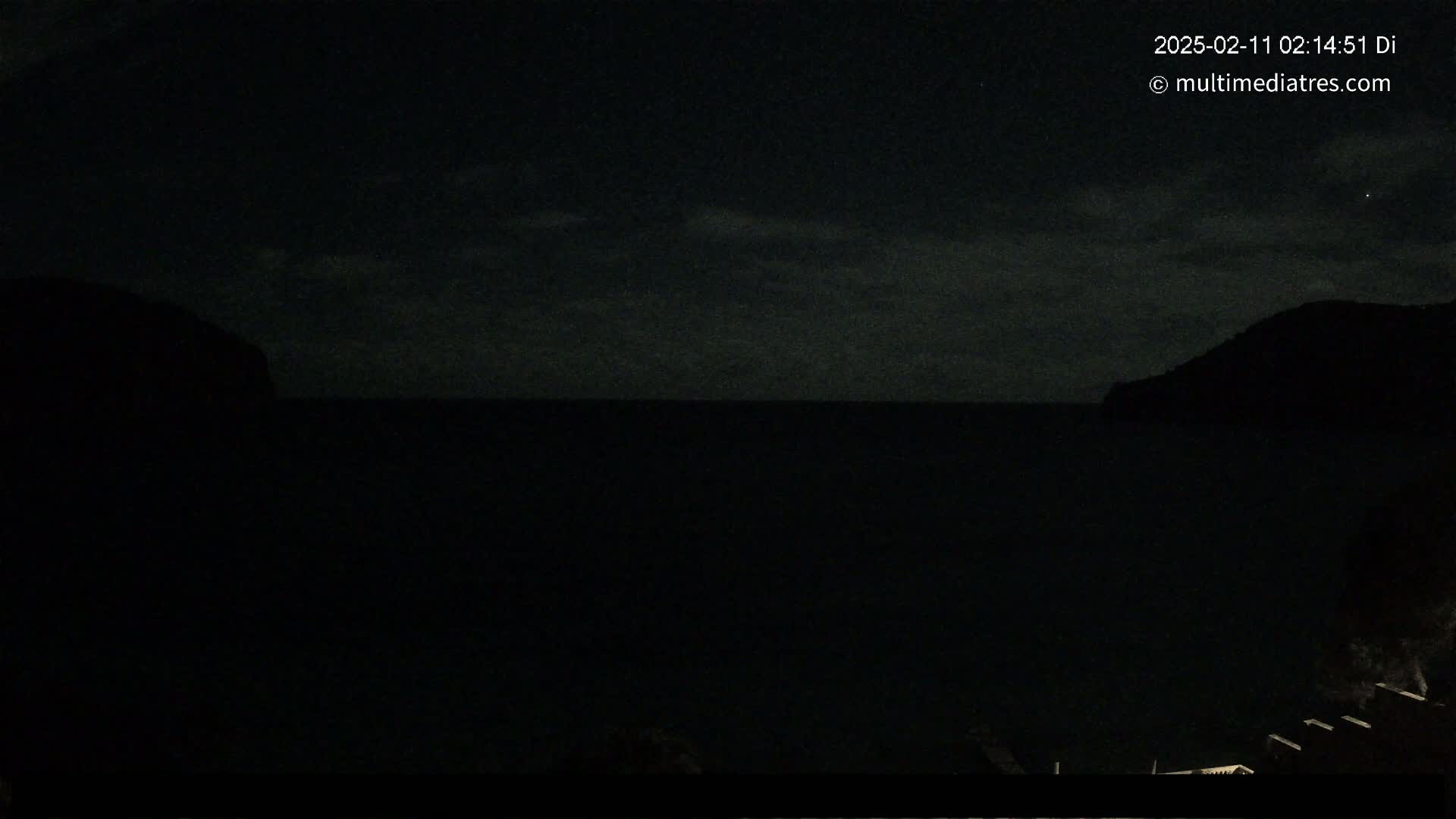 A nighttime view of a dark body of water between two silhouetted hills under a cloudy sky.