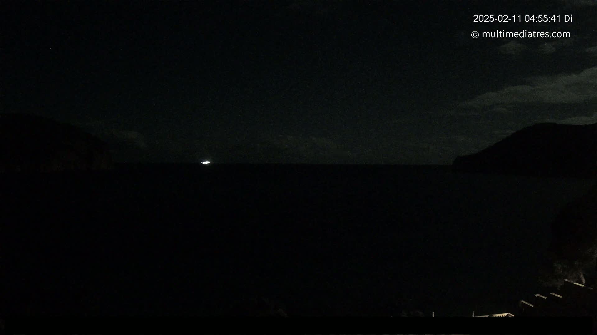 A dark, nighttime seascape shows a single, small light on the horizon between two silhouetted hills under a mostly clear sky.