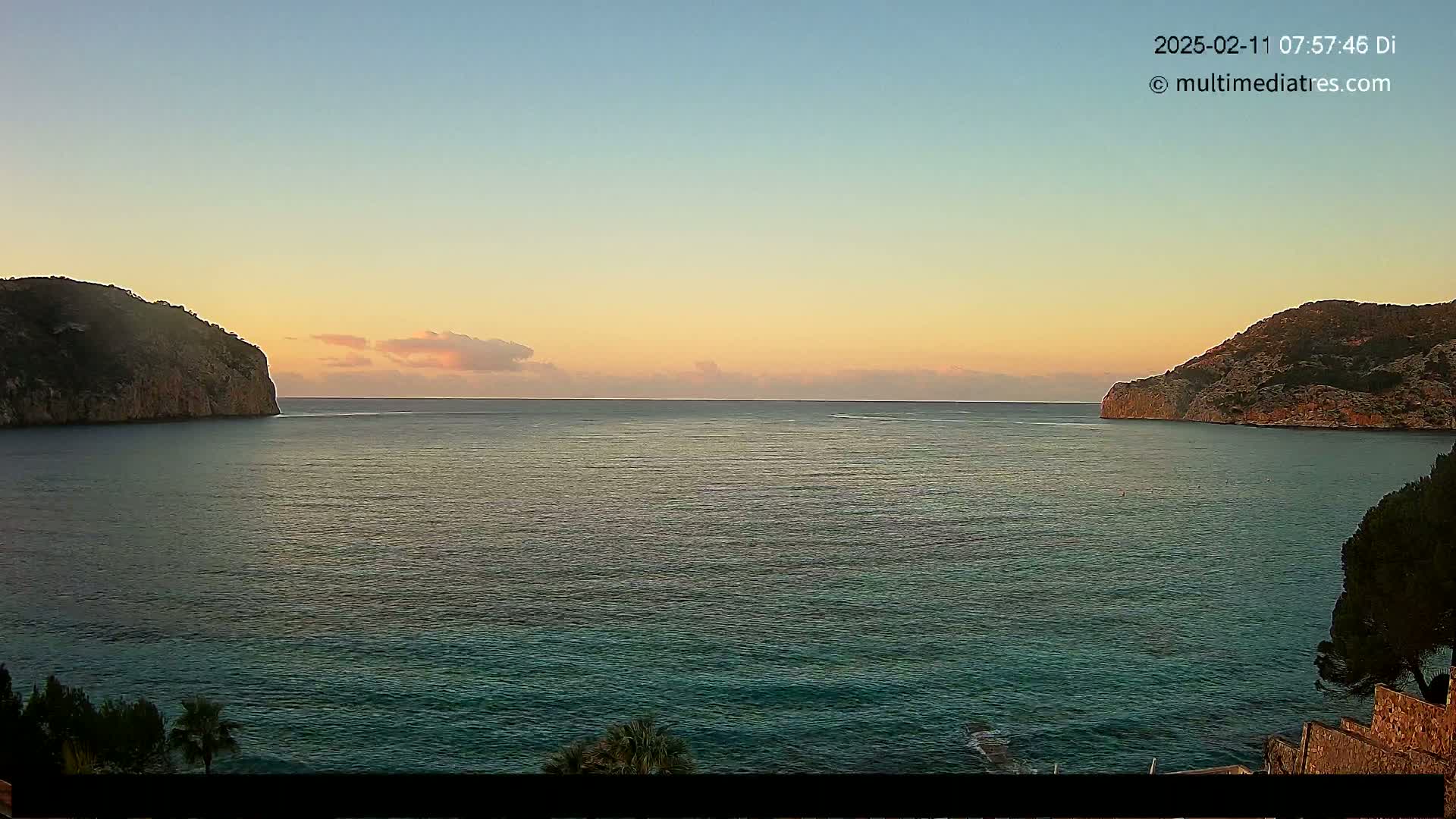 A calm sea at sunrise is nestled between two rocky headlands under a pale orange and blue sky.
