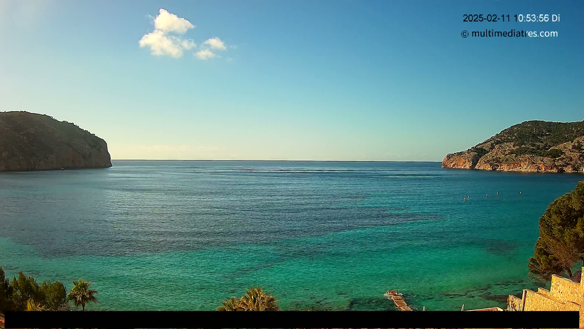 A tranquil bay of clear, turquoise water is nestled between two rocky hills under a mostly sunny sky with a single small cloud.