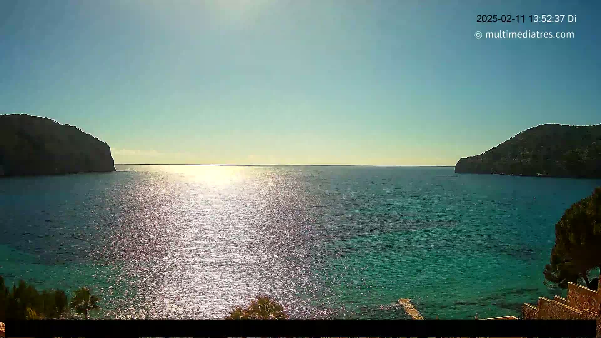 A sunny day reveals a calm, sparkling turquoise sea nestled between two dark, verdant hills.