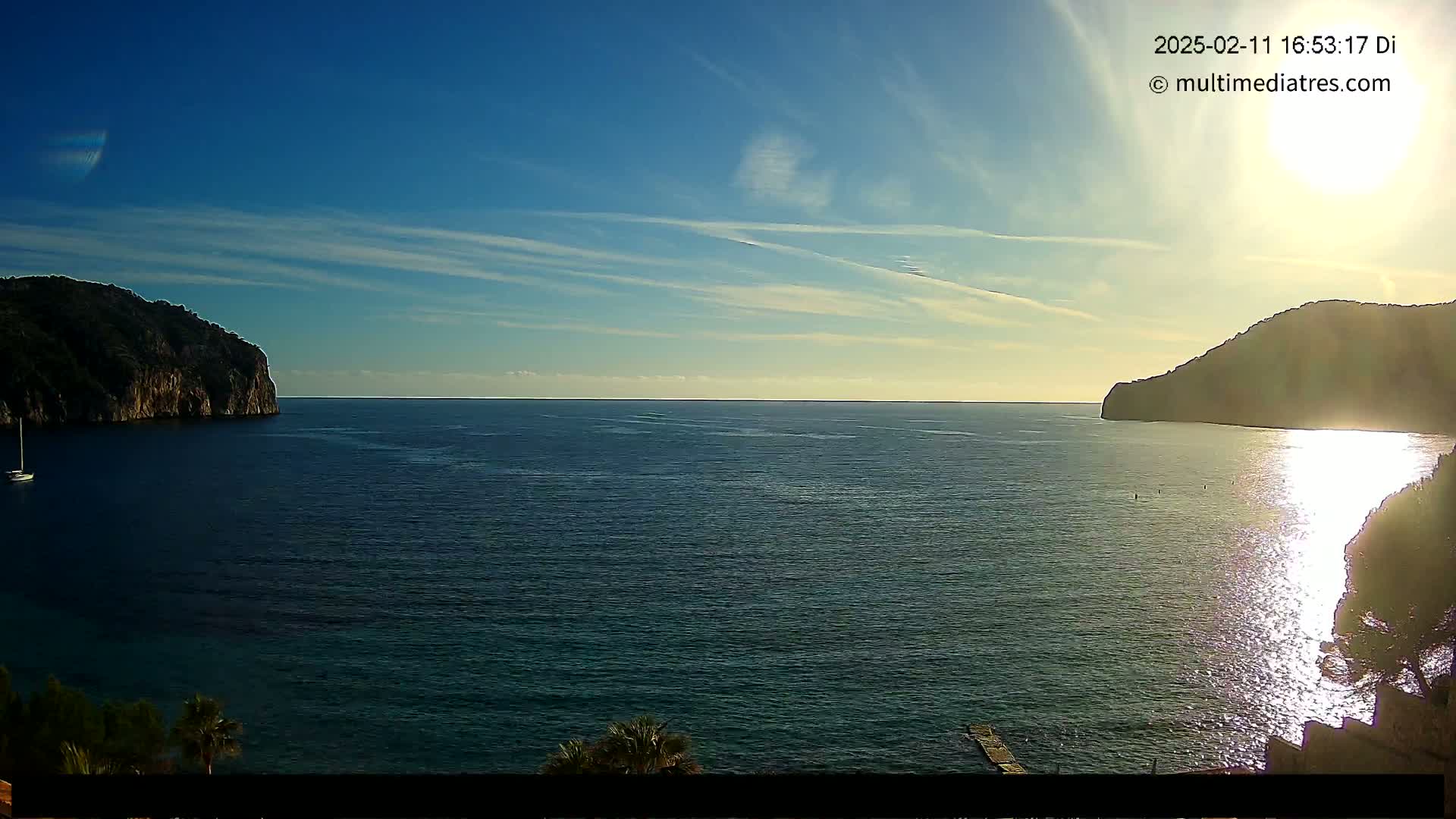 A calm, sunlit sea is seen between two dark, rocky hills under a mostly clear sky with some thin clouds.