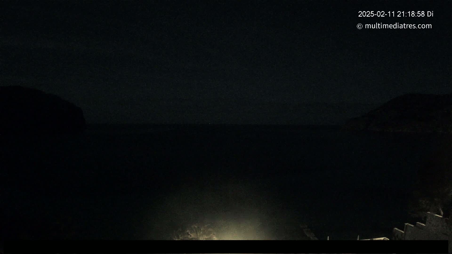 A dark, nighttime view of a calm body of water between two shadowy landmasses, illuminated only by a faint light source in the foreground.