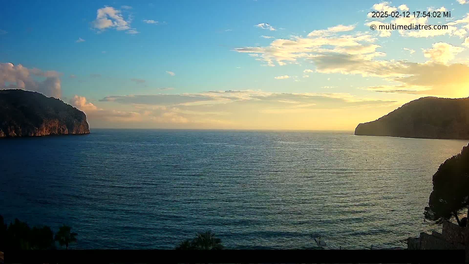 A calm sea at sunset is viewed between two dark, rocky hills under a partly cloudy sky.
