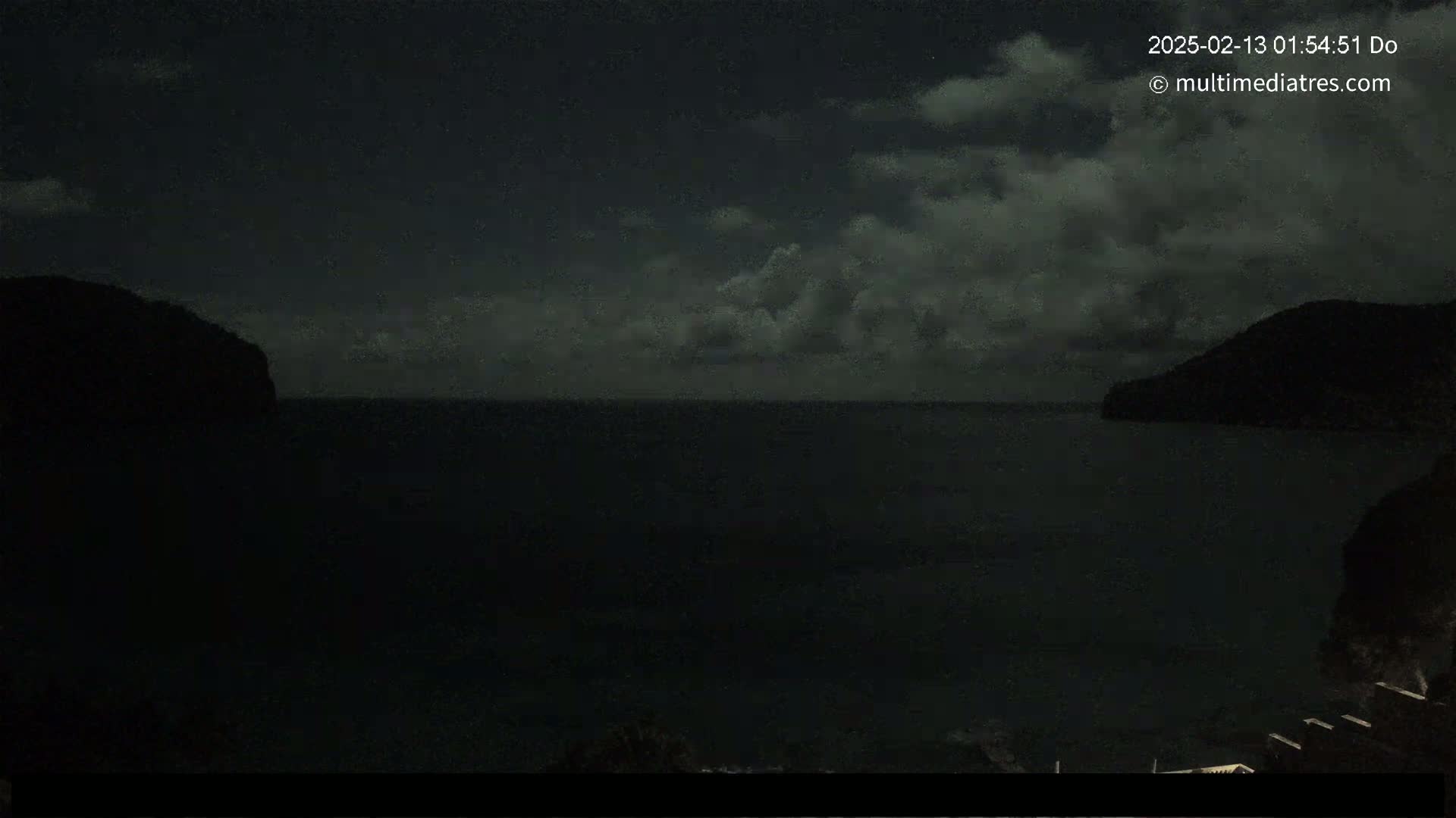 A dark, nighttime scene shows a body of water between two silhouetted hills under a cloudy sky.