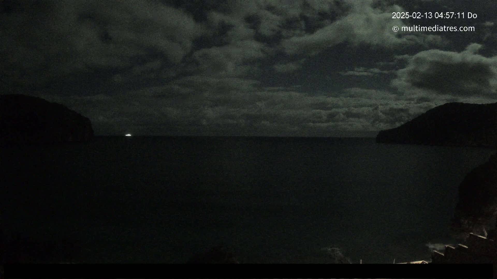 A dark, nighttime ocean scene is shown under a cloudy sky, with a distant light visible on the water between two silhouetted hills.