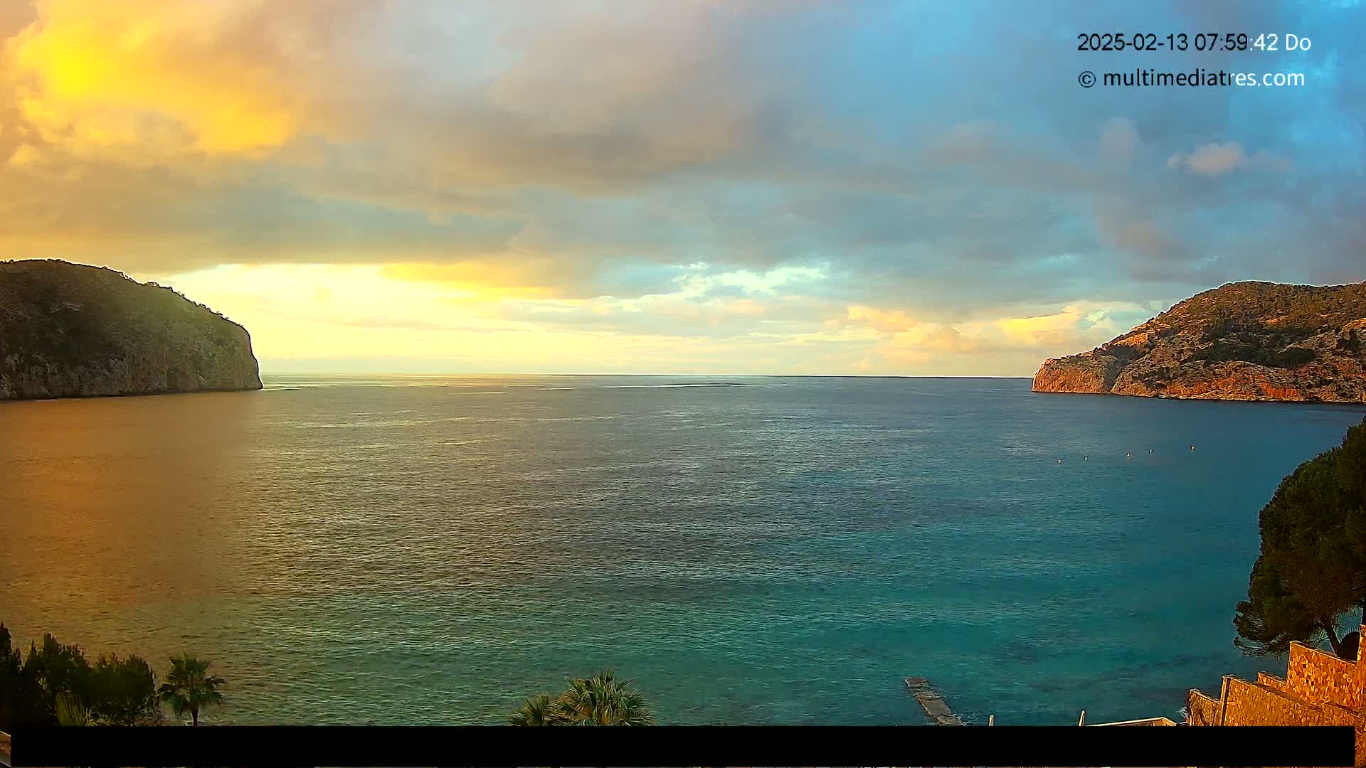 A calm sea is nestled between two rocky cliffs under a partly cloudy sunset sky.