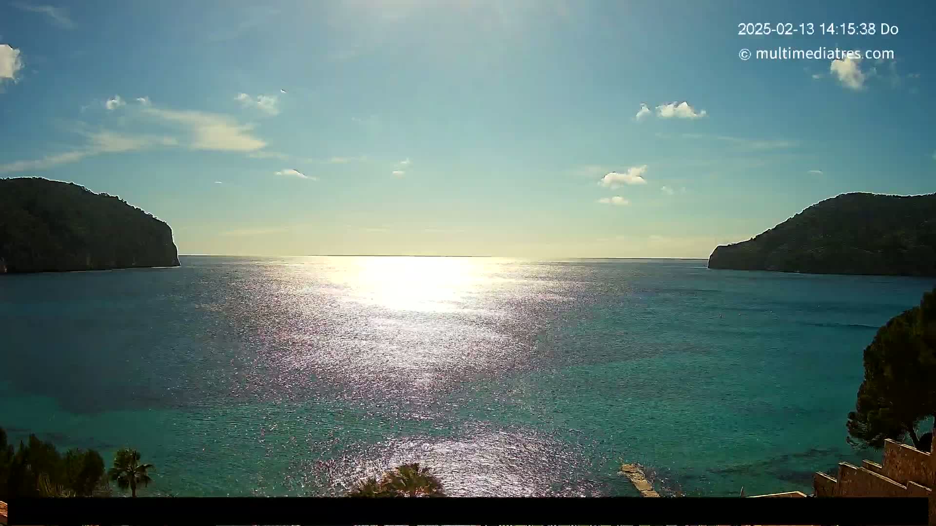 A bright, sunny day shows a calm, turquoise bay nestled between two dark, rocky hills.