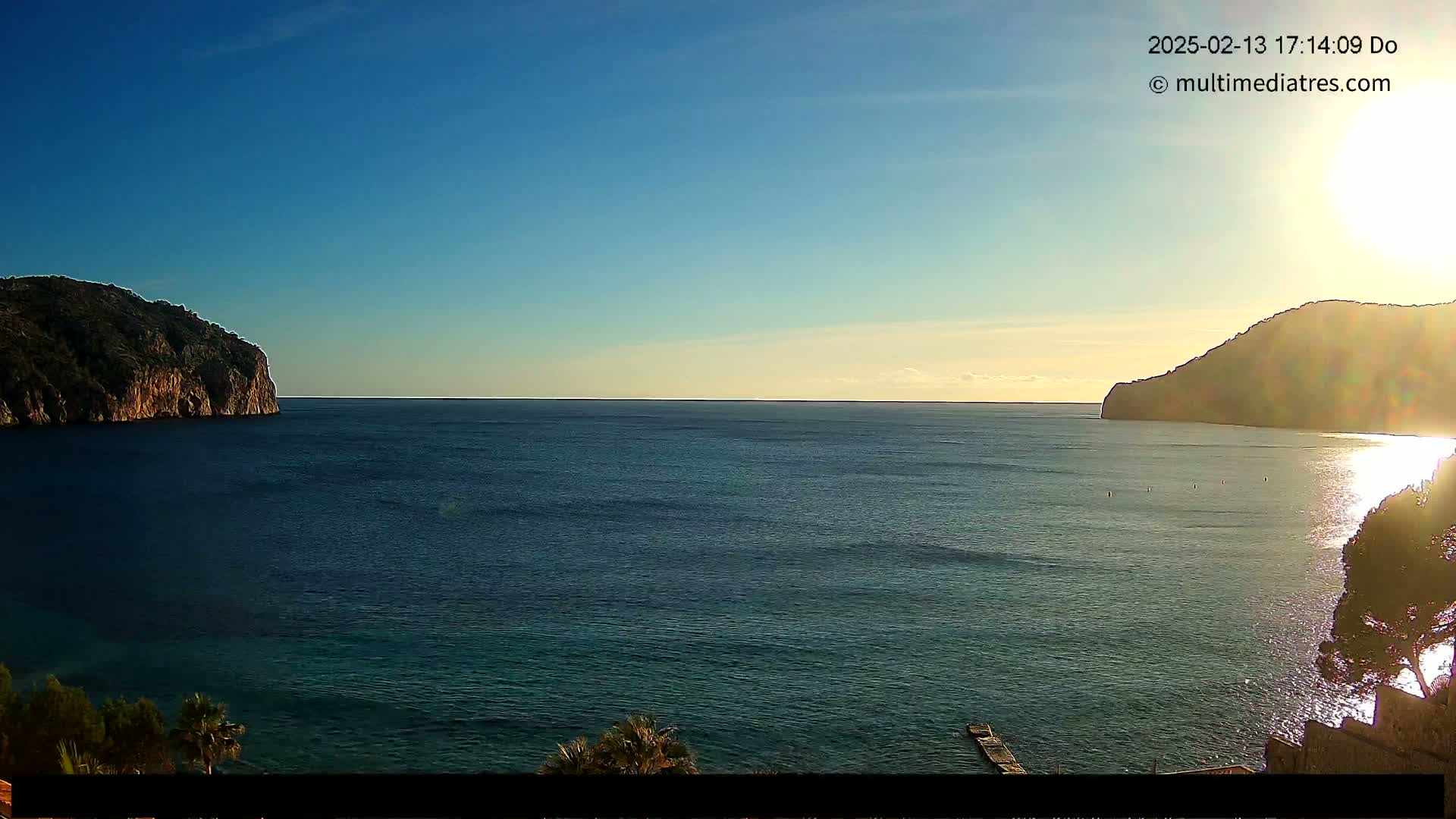 A calm, sunlit sea is seen between two dark, rocky hills under a clear blue sky.