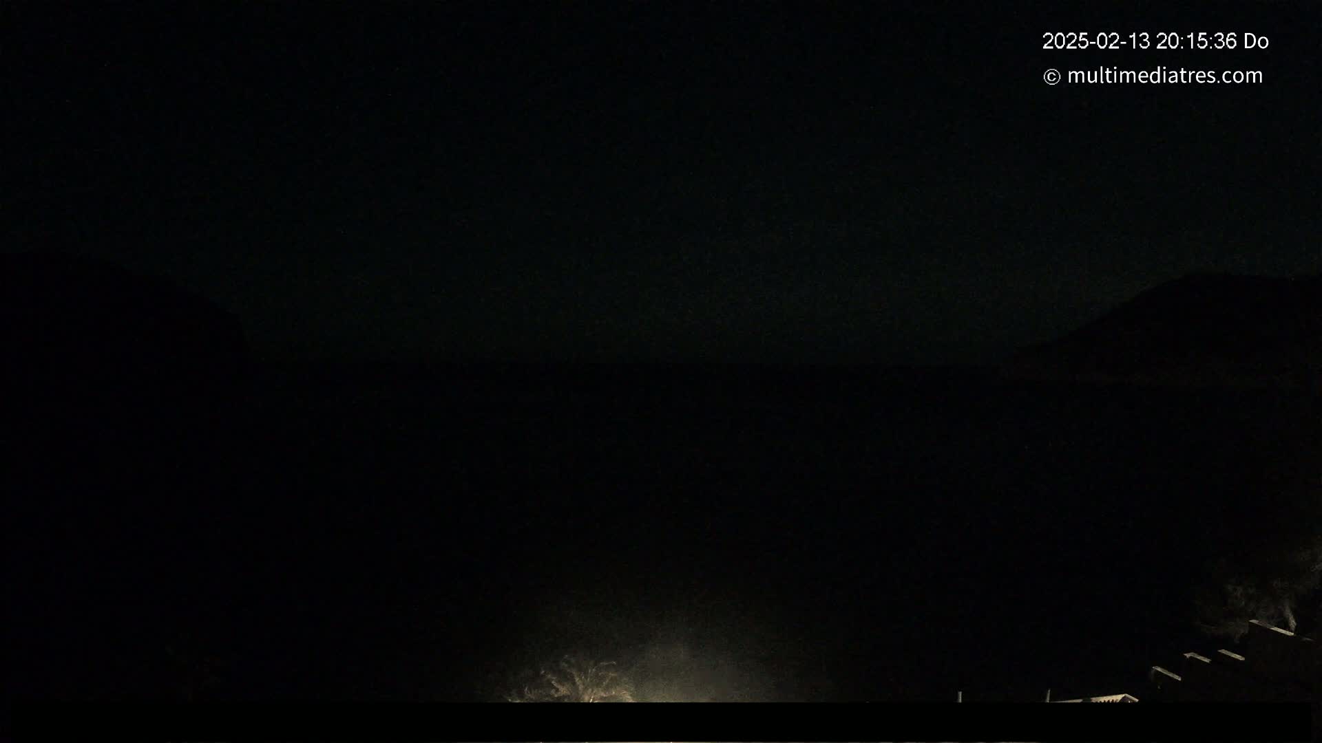 A nighttime, dark outdoor scene shows a dimly lit area near a structure, with a dark sky and indistinct land features in the background.