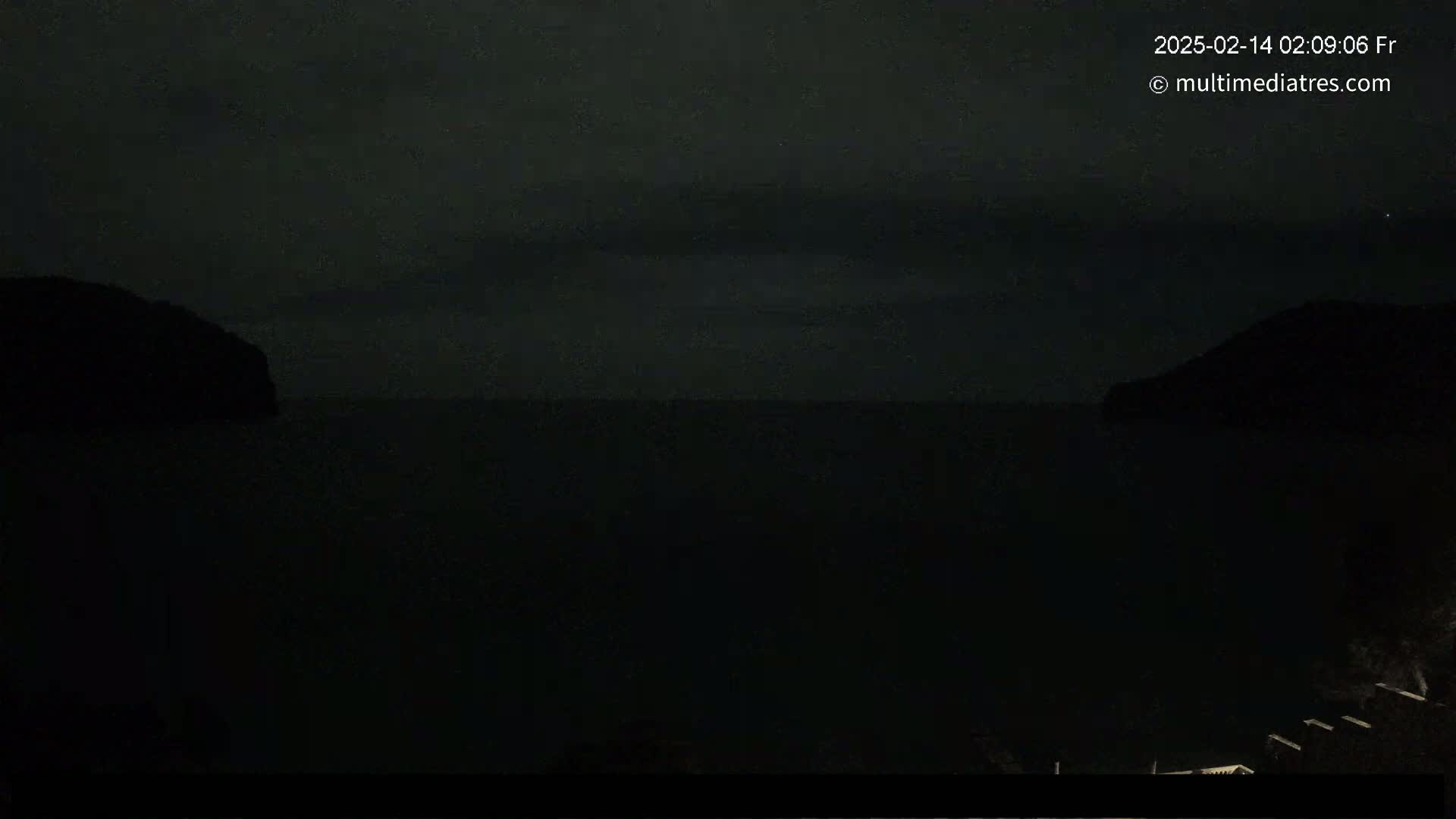 A dark, nighttime view of a calm body of water between two silhouetted hills under a cloudy sky.