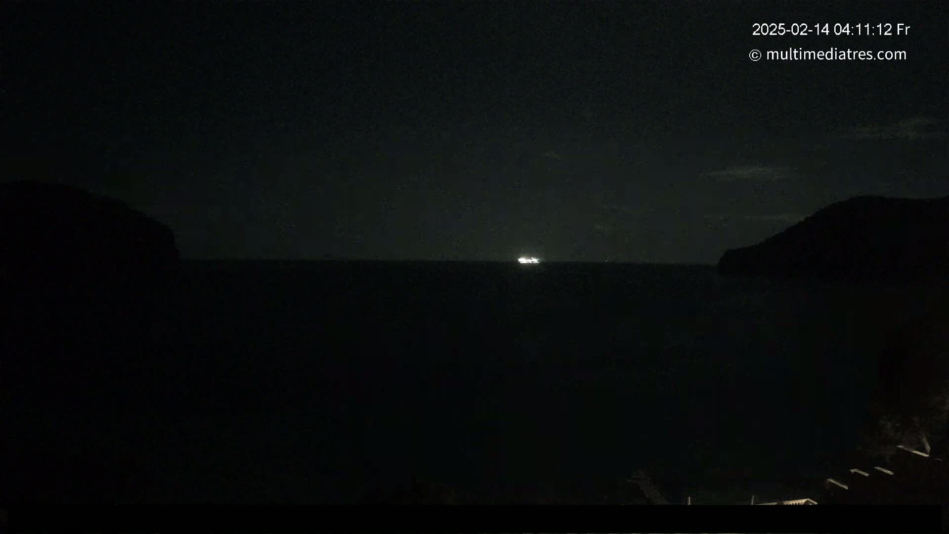A nighttime view of a dark body of water with a faintly lit ship on the horizon, flanked by silhouetted landmasses under a mostly clear sky.