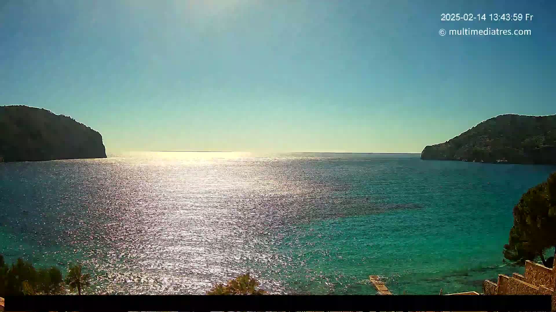 A sparkling body of turquoise water is nestled between two dark, rocky hills under a clear, sunny sky.