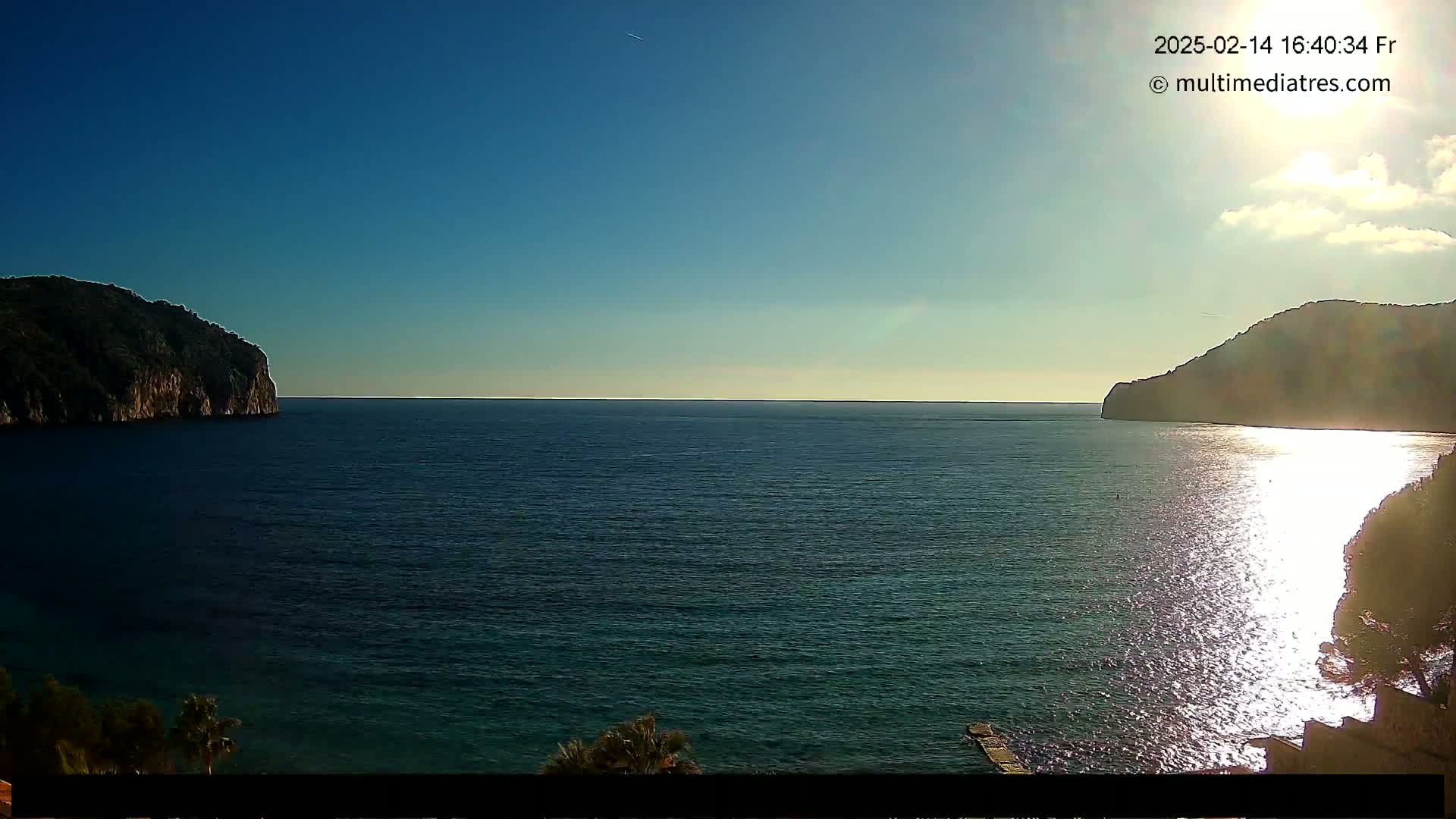 A calm, sunny day reveals a tranquil bay enclosed by dark hills, the water reflecting the sunlight.
