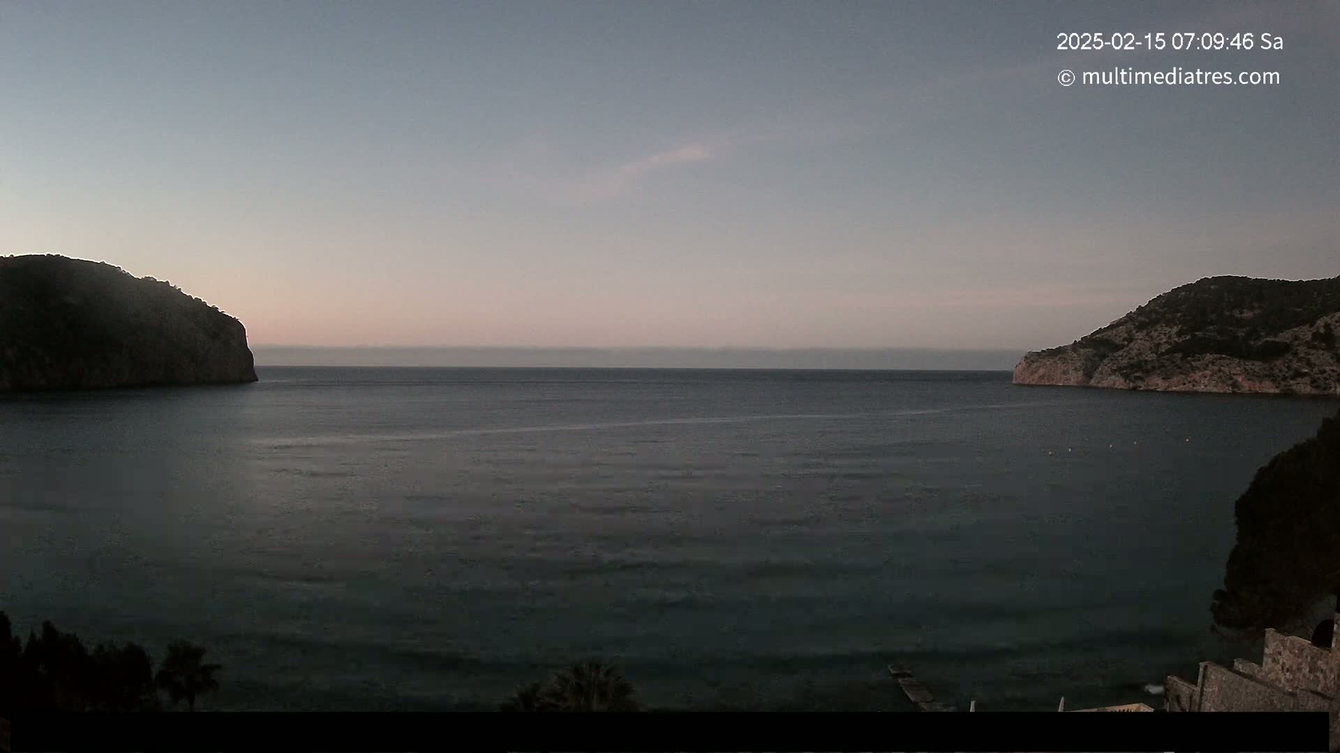 A calm, dark-blue sea is nestled between two dark, rocky hills under a pale-pink and gray twilight sky.